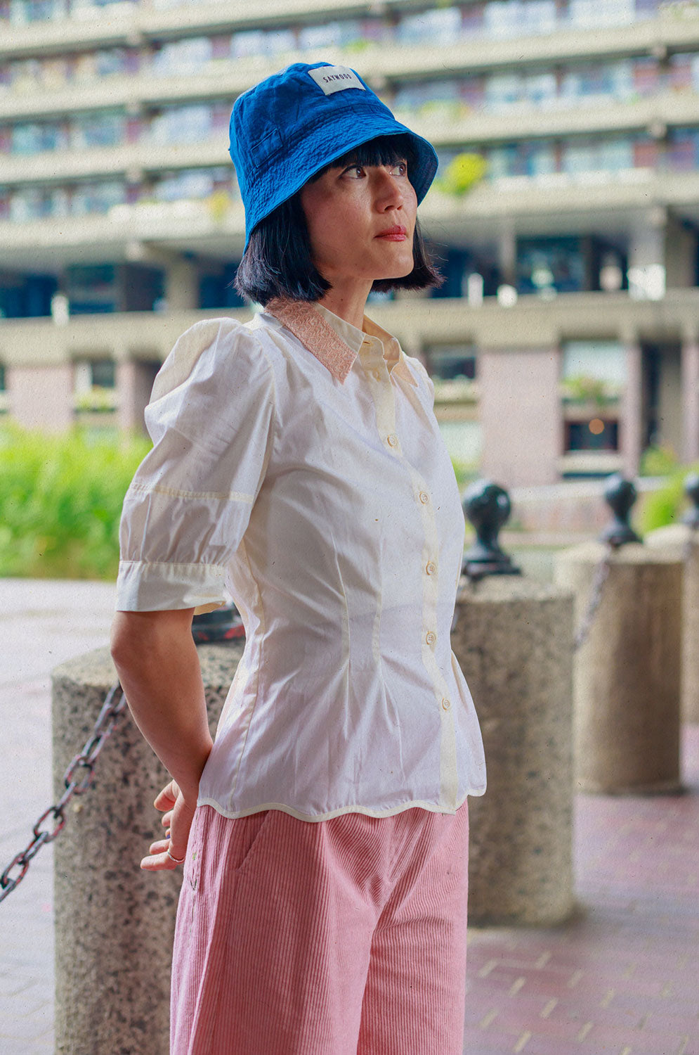 A woman of Japanese heritage stands sideways to the camera, near short posts at the Barbican centre, with her hands behind her back. She is wearing Saywood's Joni Puff Sleeve Blouse in lemon, with fit and flare shaping, peach lace collar and scalloped hem. Worn with the Amelia Wide Leg Trousers in pink cotton corduroy, the Japanese denim Bucket Hat and white sunglasses.