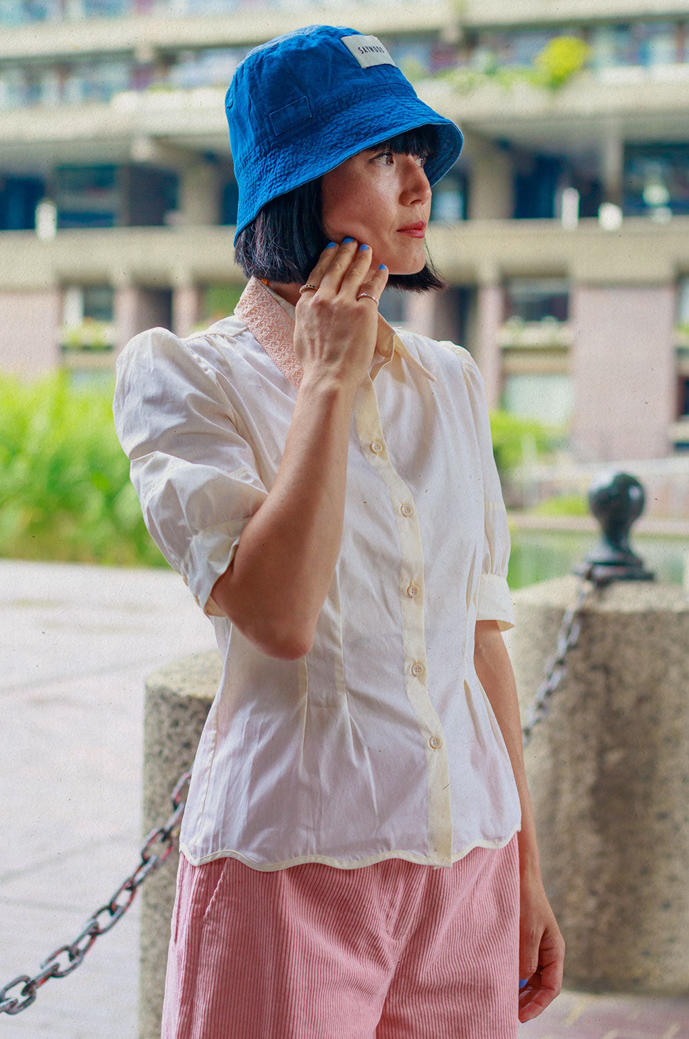 A woman of Japanese heritage stands sideways to the camera, near short posts at the Barbican centre, with one hand touching her face. She is wearing Saywood's Joni Puff Sleeve Blouse in lemon, with fit and flare shaping, peach lace collar and scalloped hem. Worn with the Amelia Wide Leg Trousers in pink cotton corduroy, the Japanese denim Bucket Hat and white sunglasses.