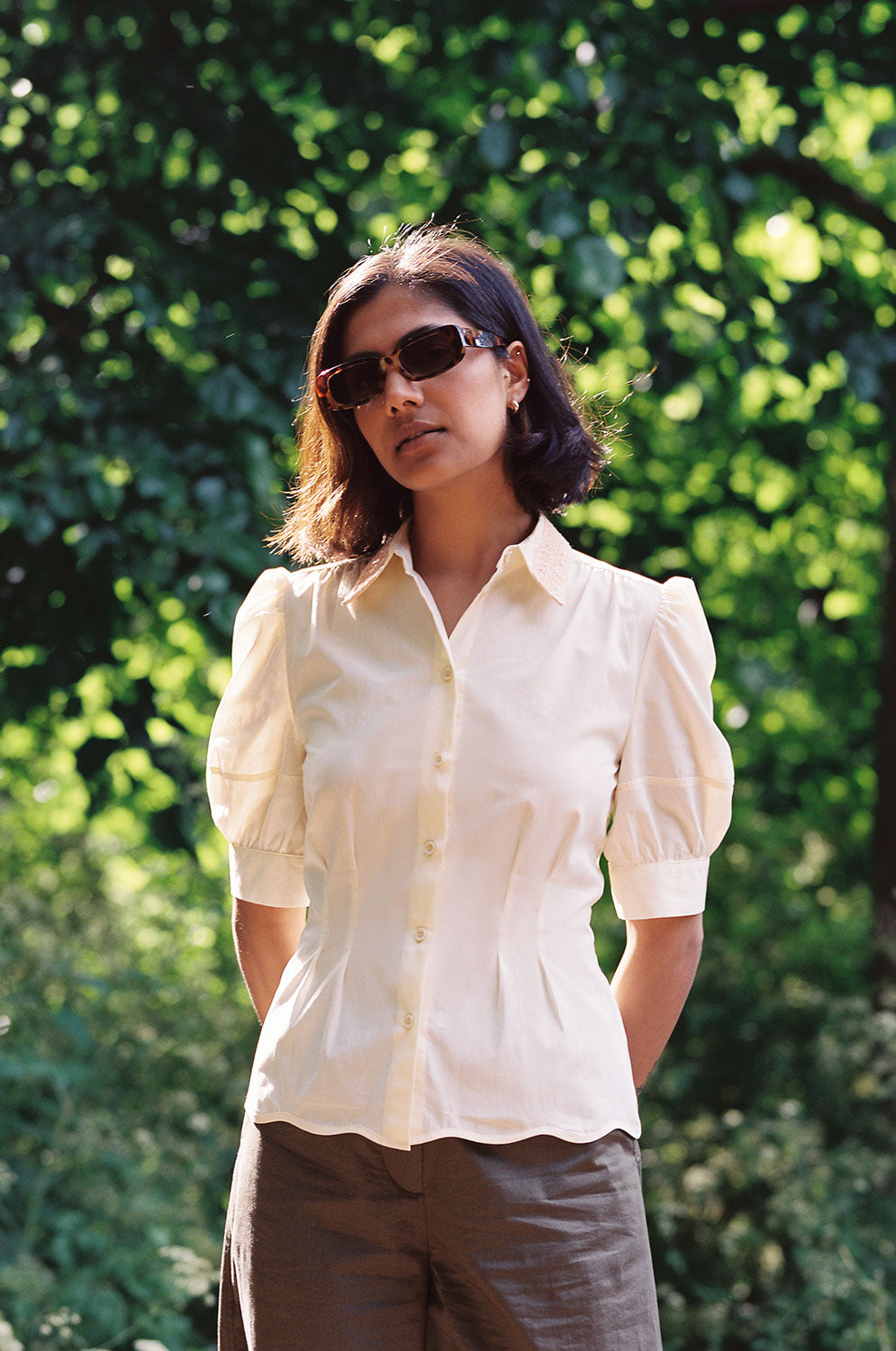 A woman stands with her hands behind her back, surrounded by nature. She is wearing Saywood's Joni Puff Sleeve Blouse, with fit and flare darting, scallop hem adn lace trim on the collar. Worn with the Amelia Wide Leg Trousers in khaki cotton, and sunglasses.