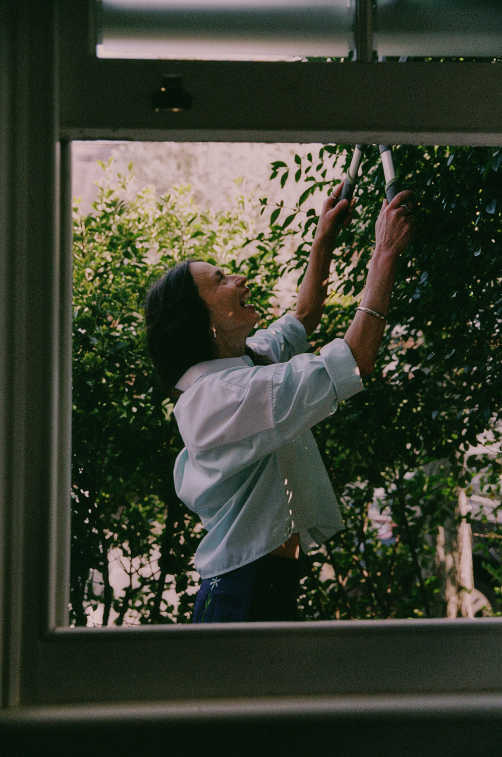 A woman is standing outside by tall green hedges, with the camera looking through an open sash window. She is holding hedge clippers and cutting the hedge. She is wearing Saywood's Lela Colourblock Boxy Shirt in green and blue pastel, with her sleeves rolled up. Worn with the Amelia Wide Leg Trousers in Japanese Denim. The signature embroidered flower can be seen on the back pocket of the trousers.