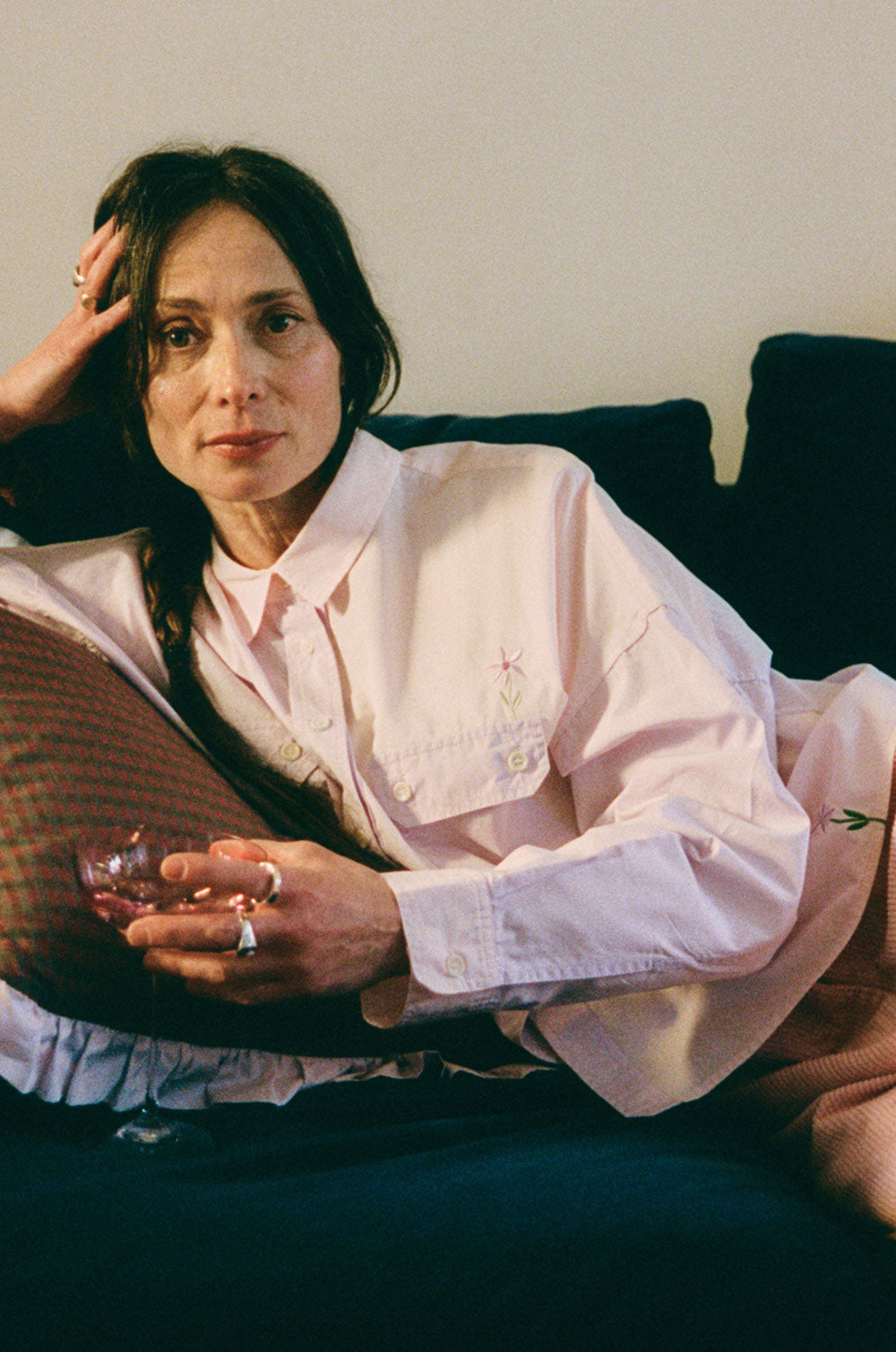 Close up image of a woman lounging on a navy sofa, leaning on a Saywood red check and lilac ruffle cushion. She is wearing Saywood's lilac and pink colourblock Jules Utility Shirt with the pink corduroy Amelia Trousers. The embroidered flower is visible on the check of the shirt above the pocket, and at the hem. She is holding a pink glass of wine with chunky silver rings on her fingers. 