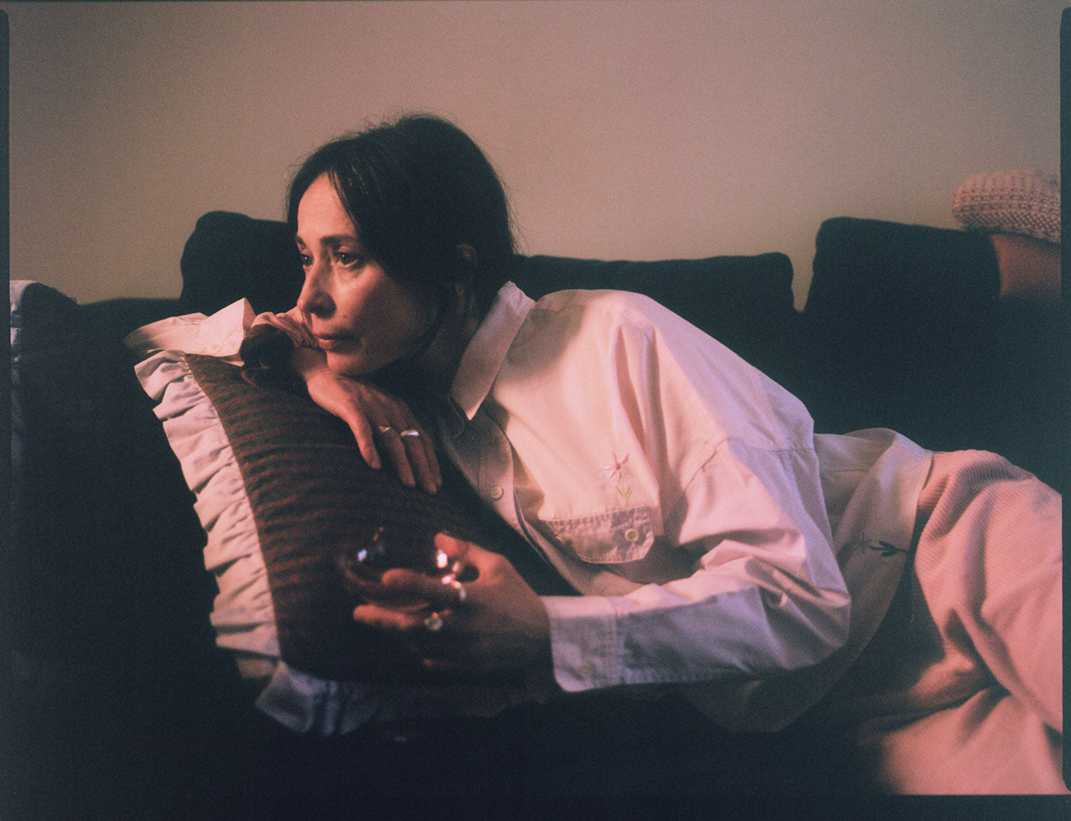 A woman is lounging on a navy sofa, leaning her chin on her hand on a Saywood red check and lilac ruffle cushion. She is wearing Saywood's lilac and pink colourblock Jules Utility Shirt with the pink corduroy Amelia Trousers. The embroidered flower is visible on the check of the shirt above the pocket, and at the hem. She is holding a pink glass of wine with chunky silver rings on her fingers. Image is shot on film.