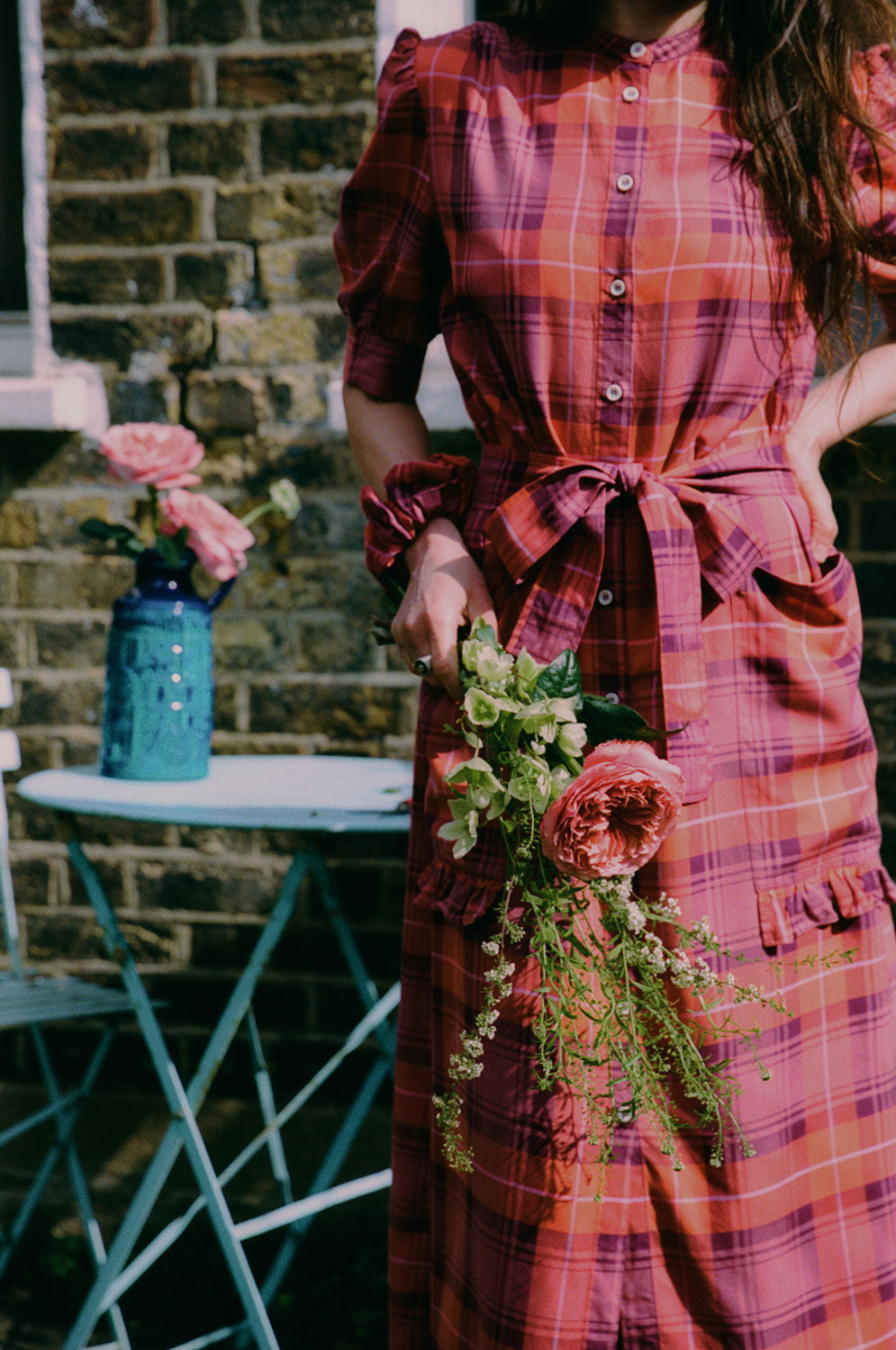 A woman stands by a blue garden table with pink flowers in a blue vintage vase on it. She is holding the same flowers in her hand and wearing Saywood's Rosa Puff Sleeve Shirtdress in coral red check with a matching scrunchie on her wrist, and matching tie belt tied round her waist. Ruffles are seen on the bottom of the dress pockets.