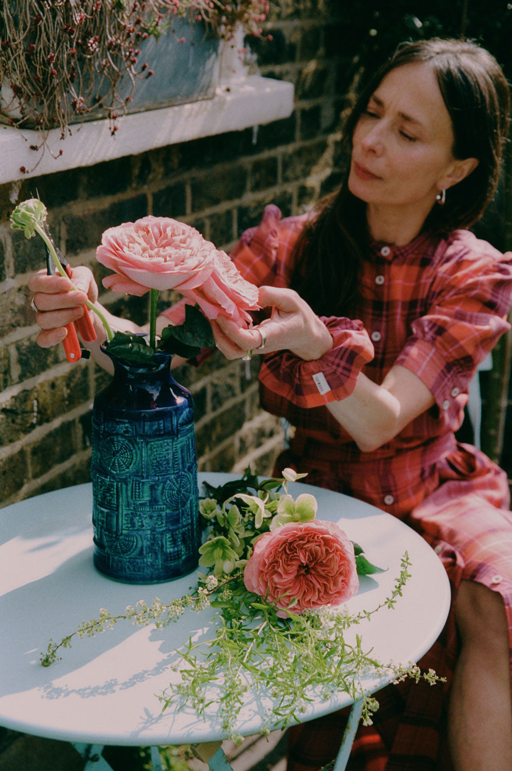 A woman sits at a blue garden table preparing pink flowers in a blue vintage vase. She is wearing Saywood's Rosa Puff Sleeve Shirtdress in coral red check with a matching scrunchie on her wrist.