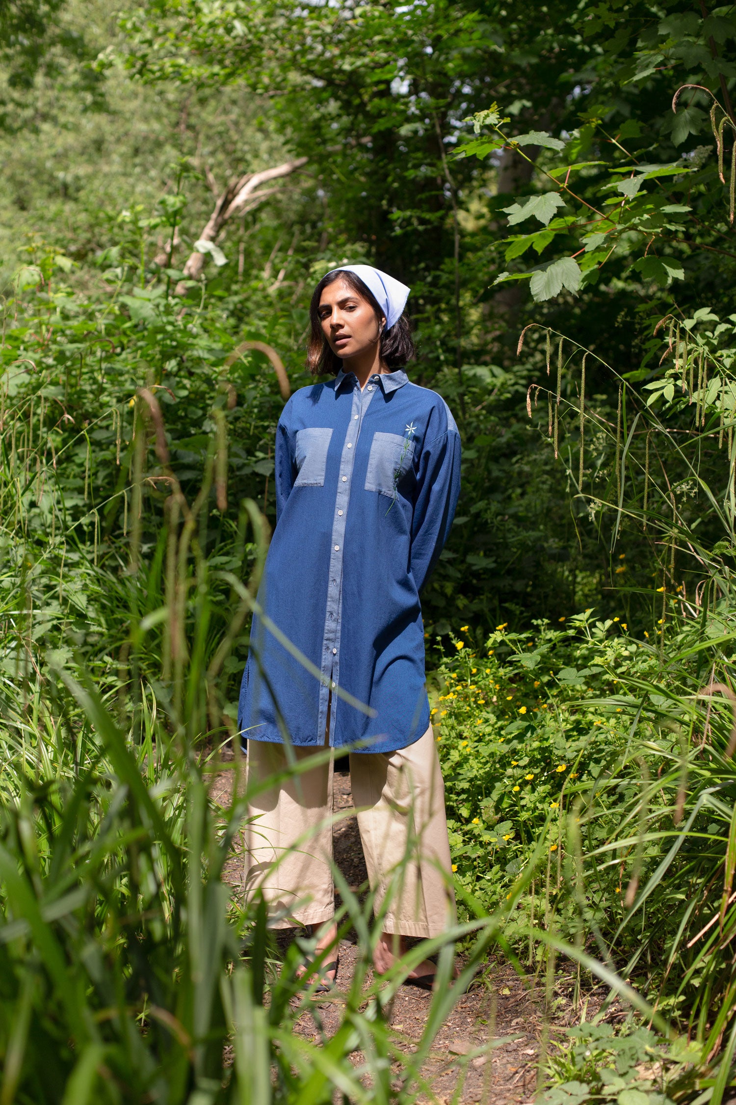 Model stands, surrounded by wild plants and greenery, with her hands behind her back. She wears Saywood's Etta Oversized Shirtdress in Japanese denim, worn over beige trousers, with a pale blue bandana in her hair made from recycled cotton.