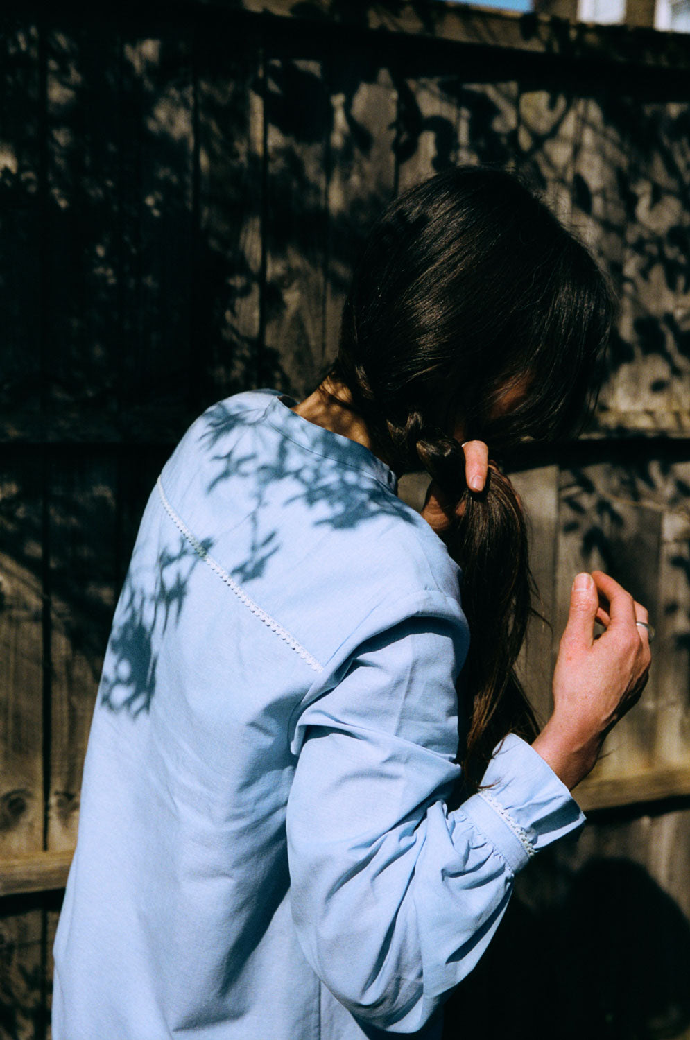 A woman stands with her back to the camera. She is wearing Saywood's Marie A-line Blouse in pale blue recycled cotton, trimmed with a narrow lace trim and gathered frill cuffs. She is plaiting her hair, whilst shadows of the trees are visible on her back. The frill of the cuffs can been seen, with lace trim on the back of the blouse at the back neck yoke.