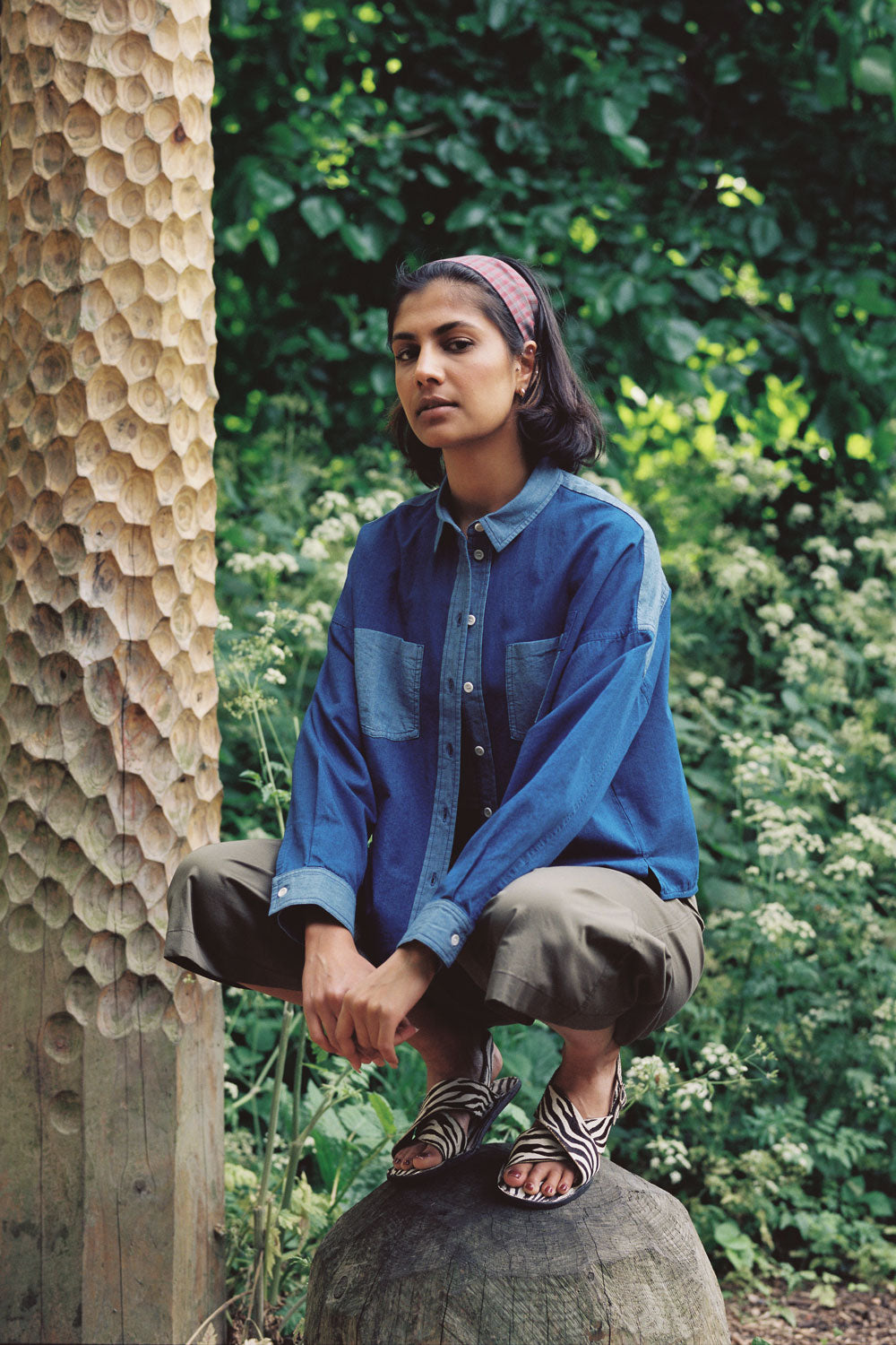 A woman squats on top of a round carved wooden bolder with a carved wooden post behind her, surrounded by greenery and nature, with her arms relaxed by her side, looking sideways. She wears Saywood's Japanese Denim Lela Patchwork Shirt, with light wash contrast cuffs, pockets and collar. Worn unbuttoned with the khaki Amelia Trousers and animal stripe monochrome sandals, with a red check headband in her hair. Shot from the front.