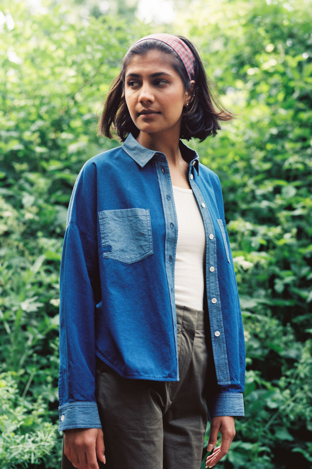 A woman stands, surrounded by greenery and nature, with her arms relaxed by her side, looking sideways. She wears Saywood's Japanese Denim Lela Patchwork Shirt, with light wash contrast cuffs, pockets and collar. Worn open over a white rib vest top with the khaki Amelia Trousers.