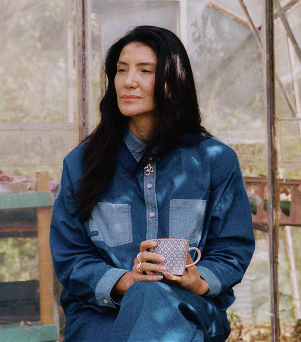 Woman holding a mug in a greenhouse setting, wearing Saywood's Lela Boxy Shirt in patchworked Japanese denim