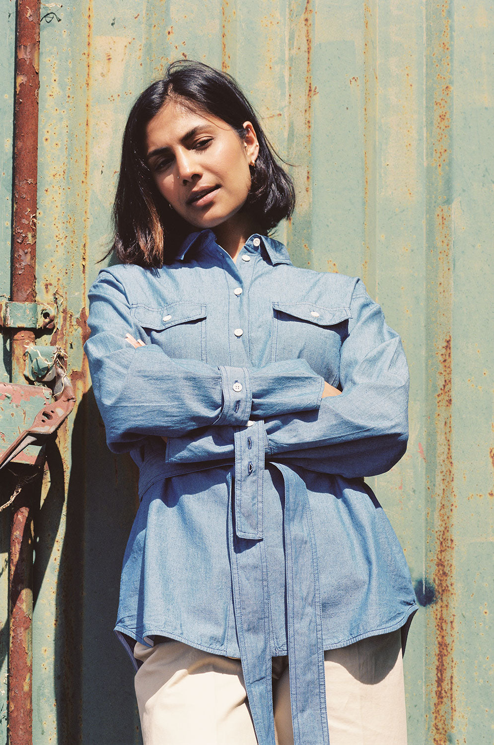 Model leans against a green shipping container, with arms crossed in front of her waist. She wears Saywood's Zadie Boyfriend Shirt in Japanese Denim with a pair of beige trousers.