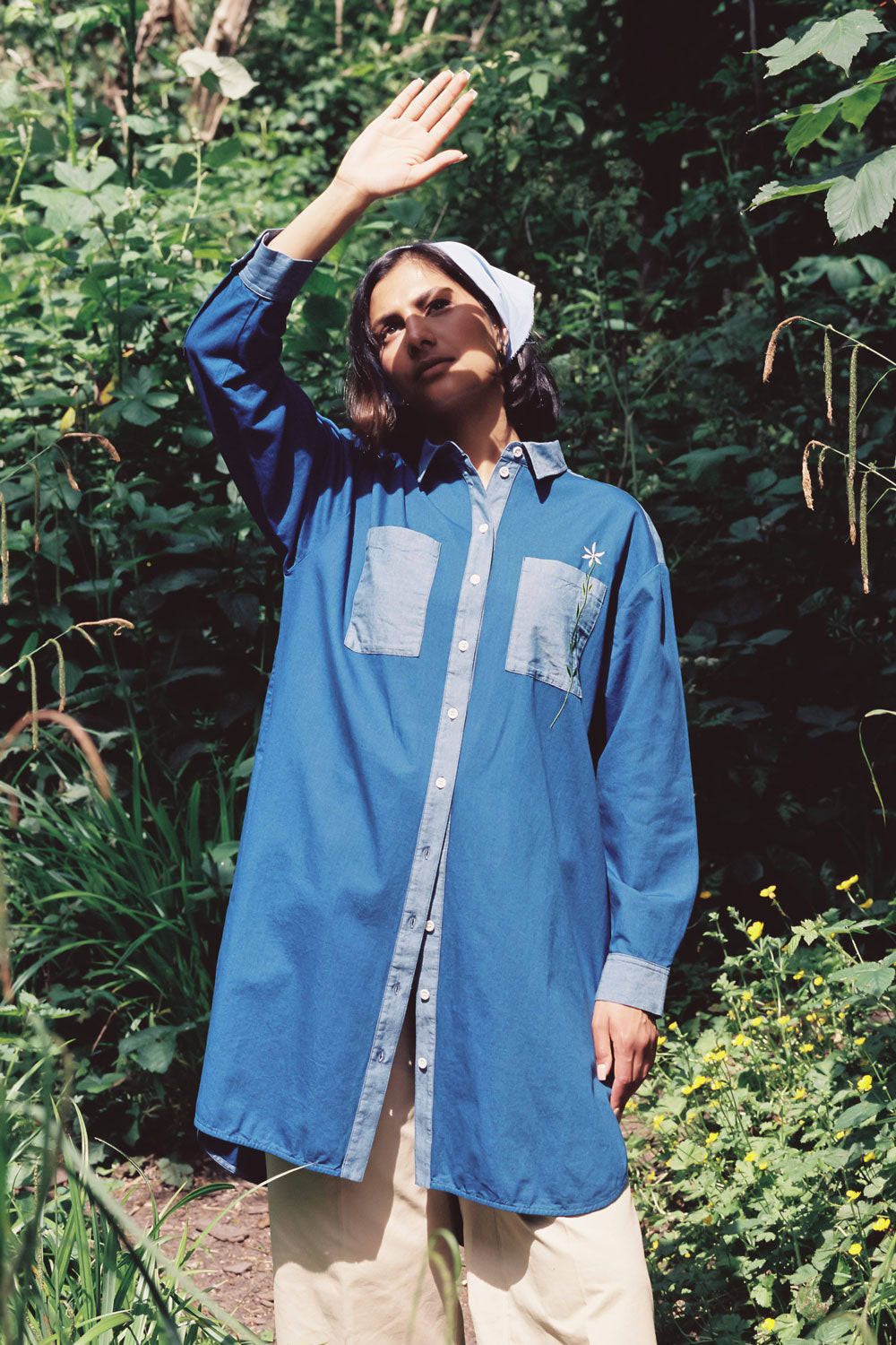 A woman stands surrounded by green nature, wearing the Etta Oversized Japanese Denim Shirtdress, worn over tan trousers, with a blue bandana in recycled cotton. One hand is held up to the sky to block the sunlight.