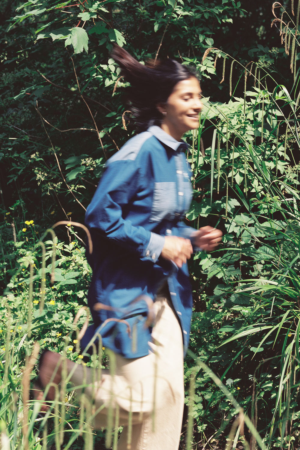 A woman is running through green nature, wearing the Etta Oversized Japanese Denim Shirtdress, worn over tan trousers.