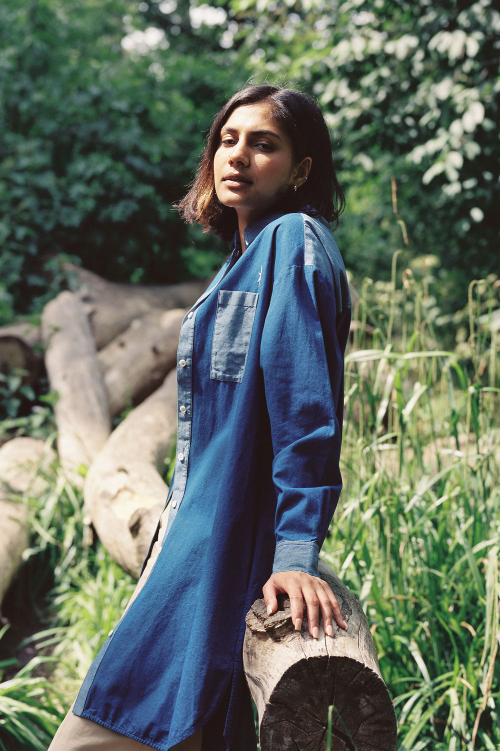 A woman sits on a log, shot from the side, surrounded by green nature, looking towards the camera. She is wearing the Etta Oversized Japanese Denim Shirtdress, worn over tan trousers.