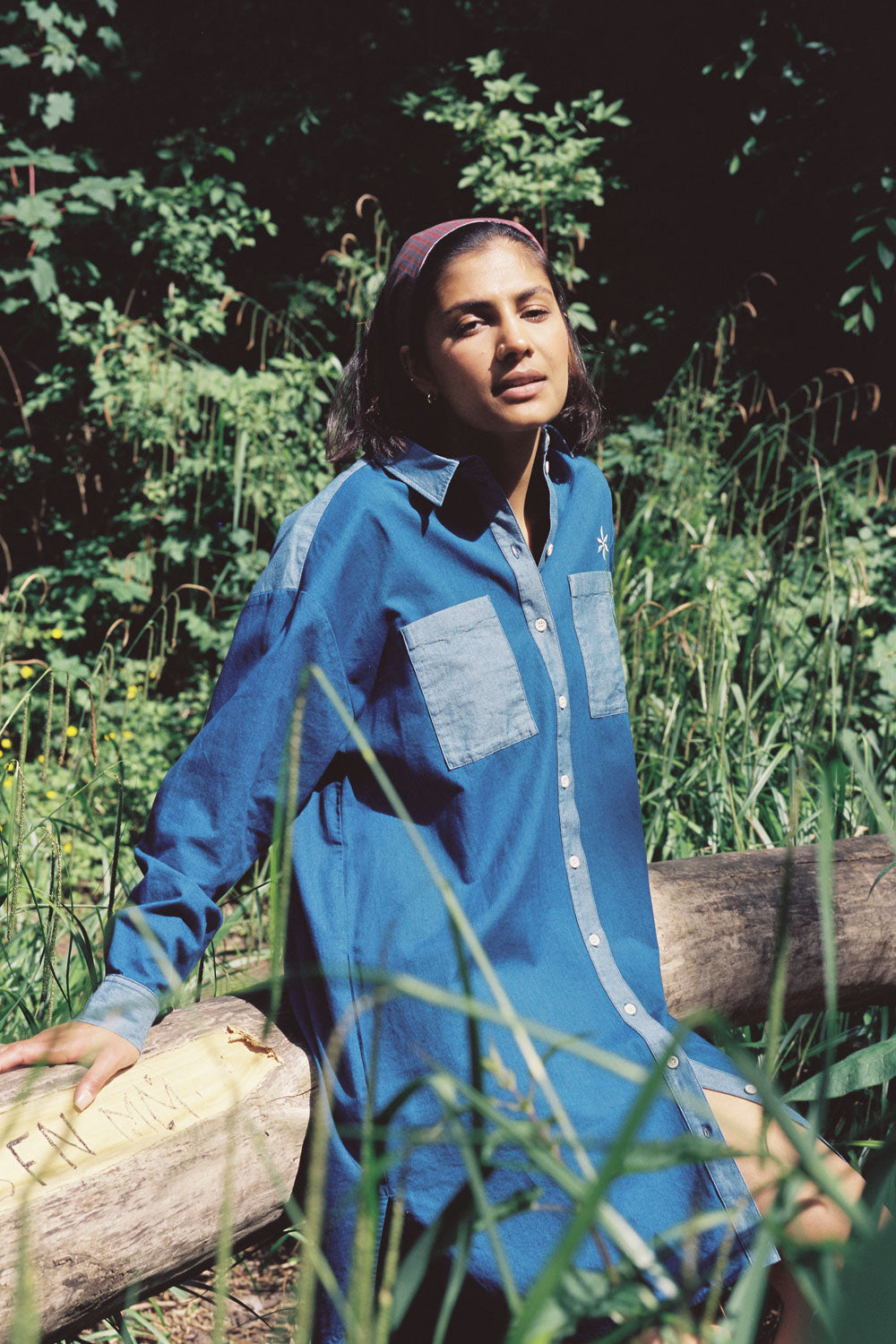 A woman sits on a log, surrounded by green nature, looking towards the camera. She is wearing the Etta Oversized Japanese Denim Shirtdress, with a red check headband in her hair.