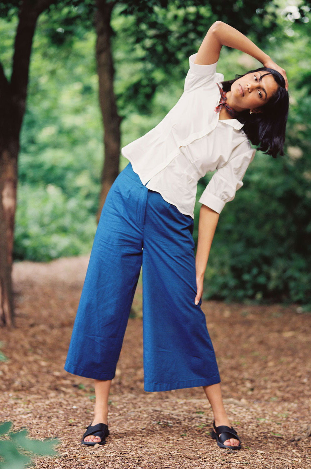 A woman stands out in nature surrounded by trees. She stands with her legs a part leaning bent to one side with one hand on her head. She wears Saywood's Amelia Wide Leg Culotte Trousers in Japanese denim with the Joni fit and flare blouse and scalloped hem in white. Worn with sandals and a red check neck tie.