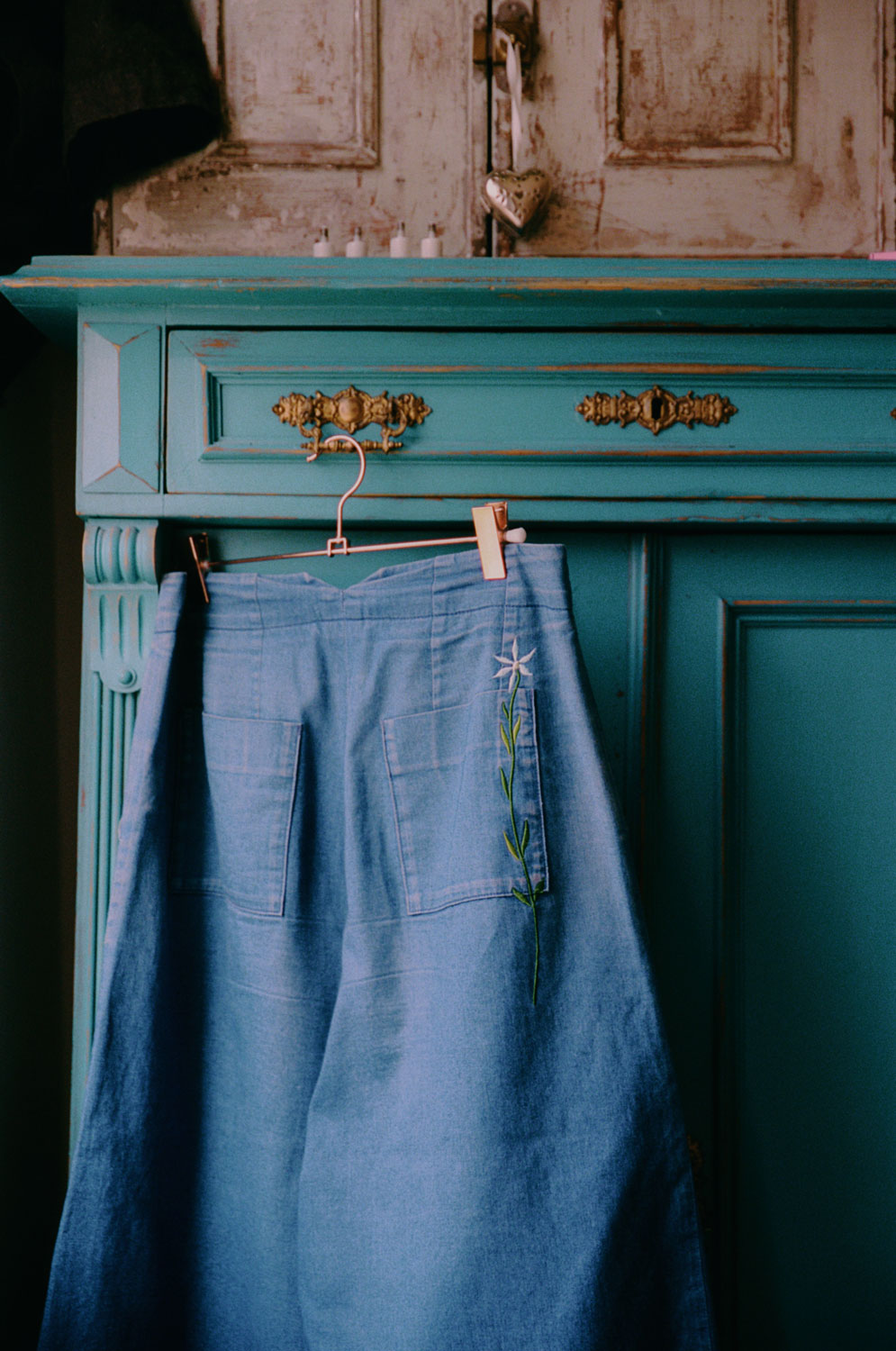 The Japanese denim Amelia Wide Leg Culotte Trousers are hanging on a rose gold hanger from the ornate gold handle of a turquoise vintage wooden cupboard, with metal cabinet on top. The back of the trousers can be seen, with the signature Saywood embroidered flower on the back pocket, and triangle cut out at the waist. The trousers have been worn so the denim has slightly washed down with age.