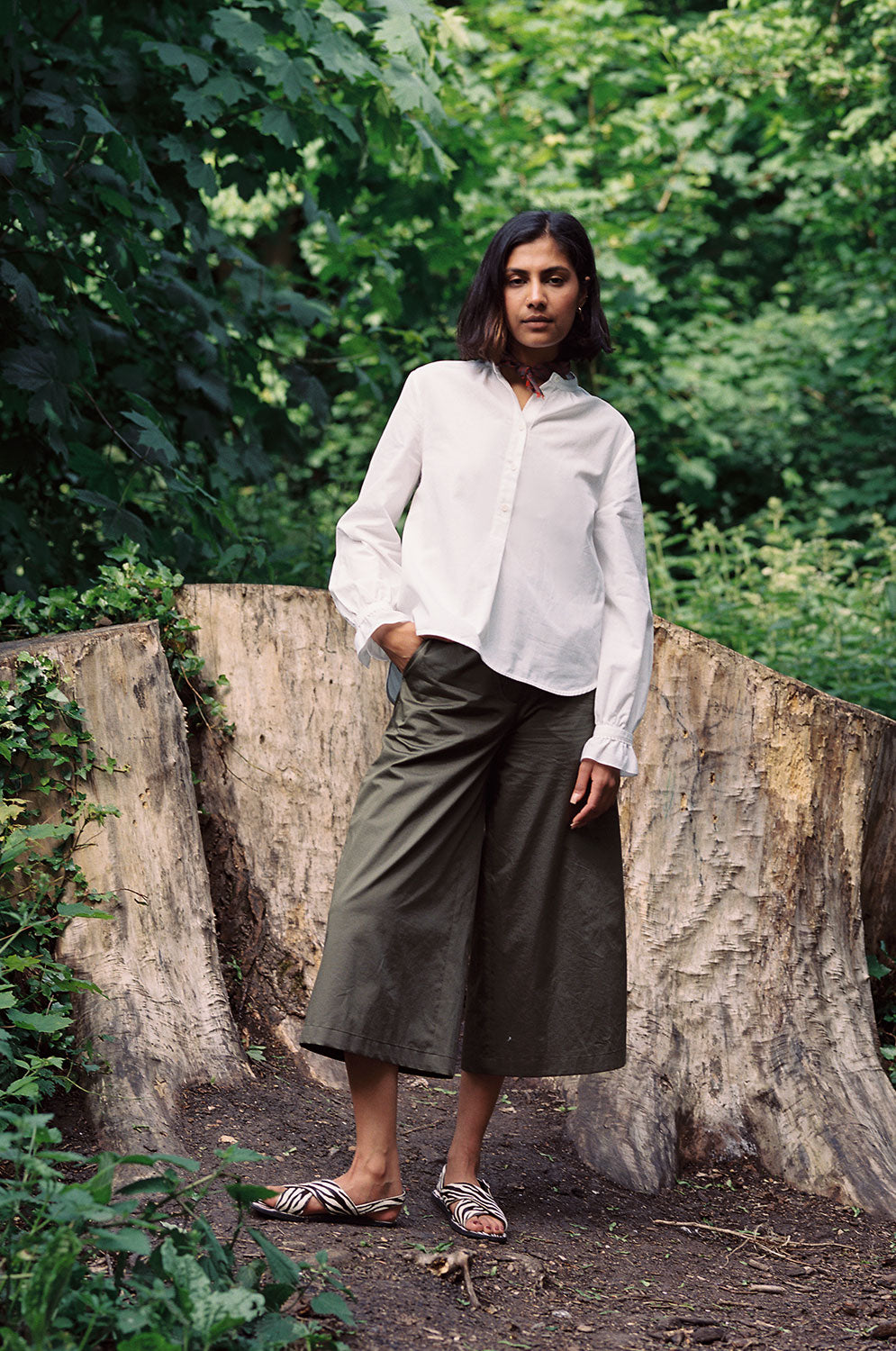 Model stands, one knee bent, in front of tree stumps and greenery. She wears Saywood's Amelia wide leg trouser in khaki cotton sateen, with the white Marie A-line Blouse with a red check neckerchief tied round her neck. Worn with black and white zebra print sandals.