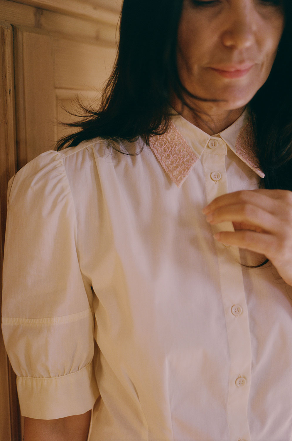 Close up of a woman with dark hair, standing in front of a wooden wardrobe, wearing Saywood's Joni Puff Sleeve Blouse in lemon, soft pale yellow. The gathered puff sleeves, gathered shoulders and orange lace collar can be seen.