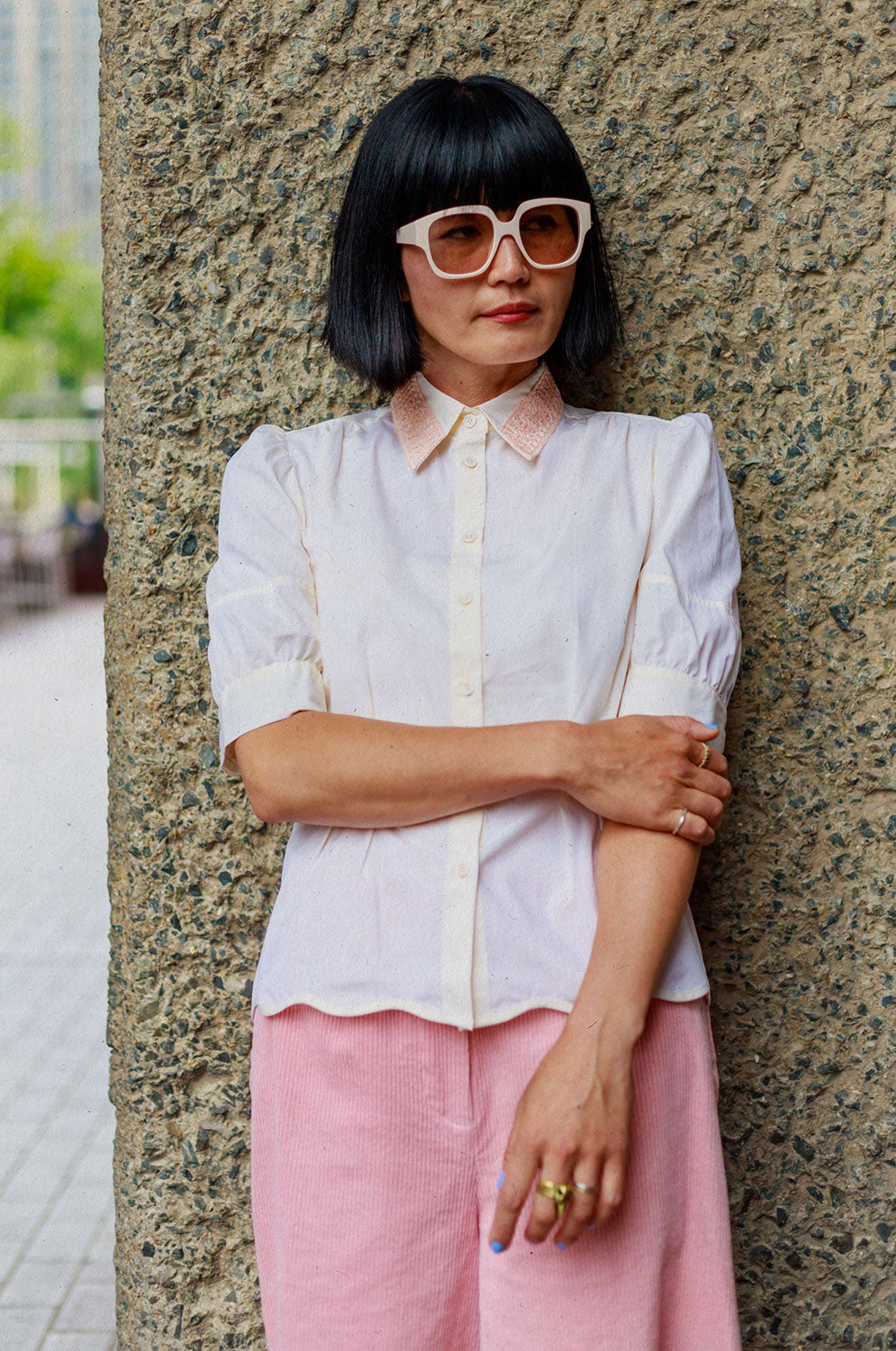 A woman of Japanese heritage leans her back against a rough concrete wall, looking slightly to the side, with one hand holding her elbow across her body. She is wearing Saywood's Joni Puff Sleeve Blouse in lemon, with fit and flare shaping, peach lace collar and scalloped hem. Worn with the Amelia Wide Leg Trousers in pink cotton corduroy and white sunglasses.