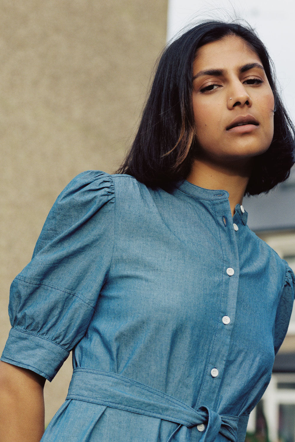 Close up of a woman wearing Saywood's Rosa denim shirtdress in light wash Japanese denim; a long line style with patch pockets and ruffles, a belt tied round the waist. The gathered puff sleeve can be see as a focal point in the shot.