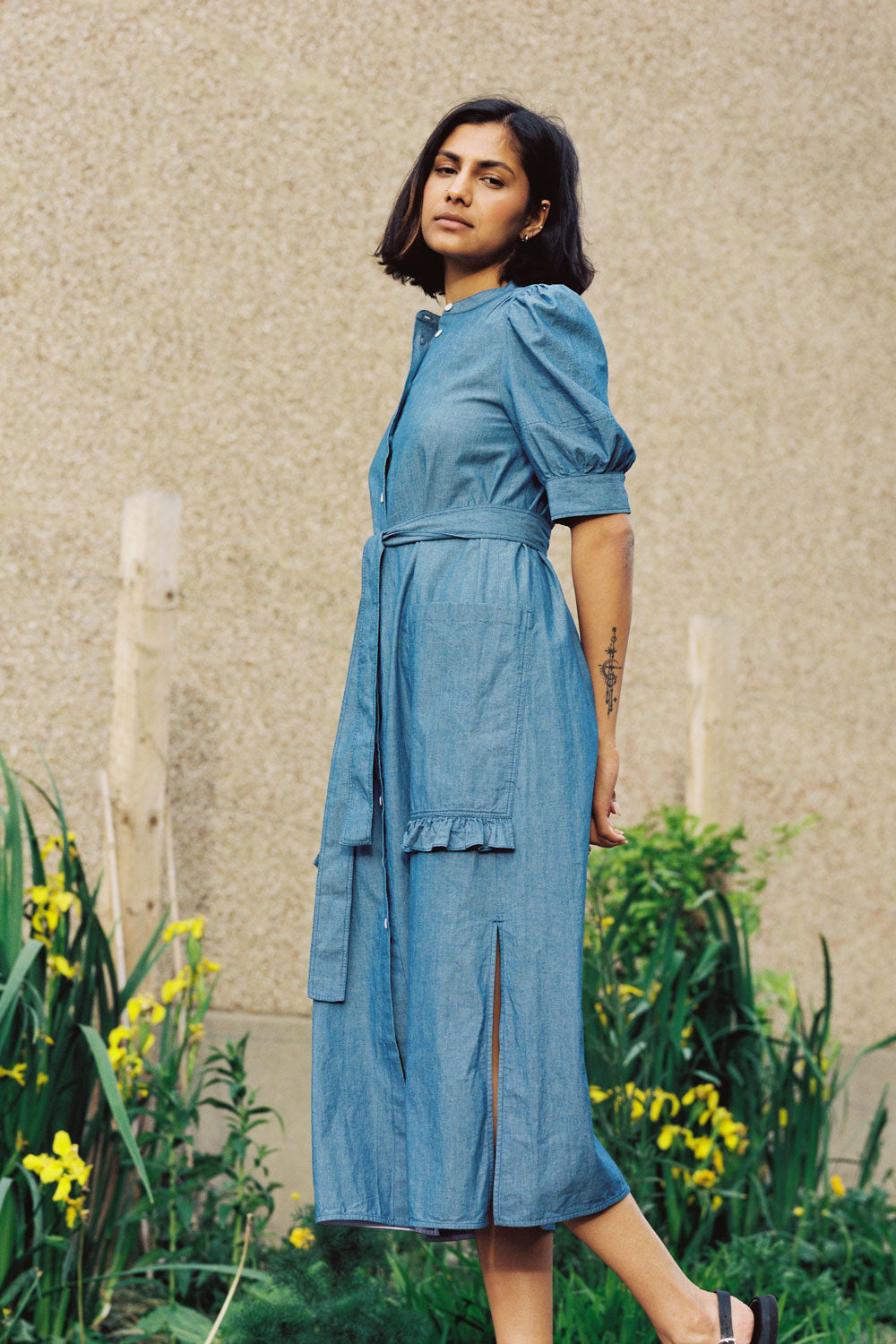 A woman stands wearing Saywood's Rosa denim shirtdress in light wash Japanese denim; a long line style with patch pockets and ruffles, a belt tied round the waist. She stands with her hands behind her back, turned towards the side. Yellow flowers can be seen in the background.