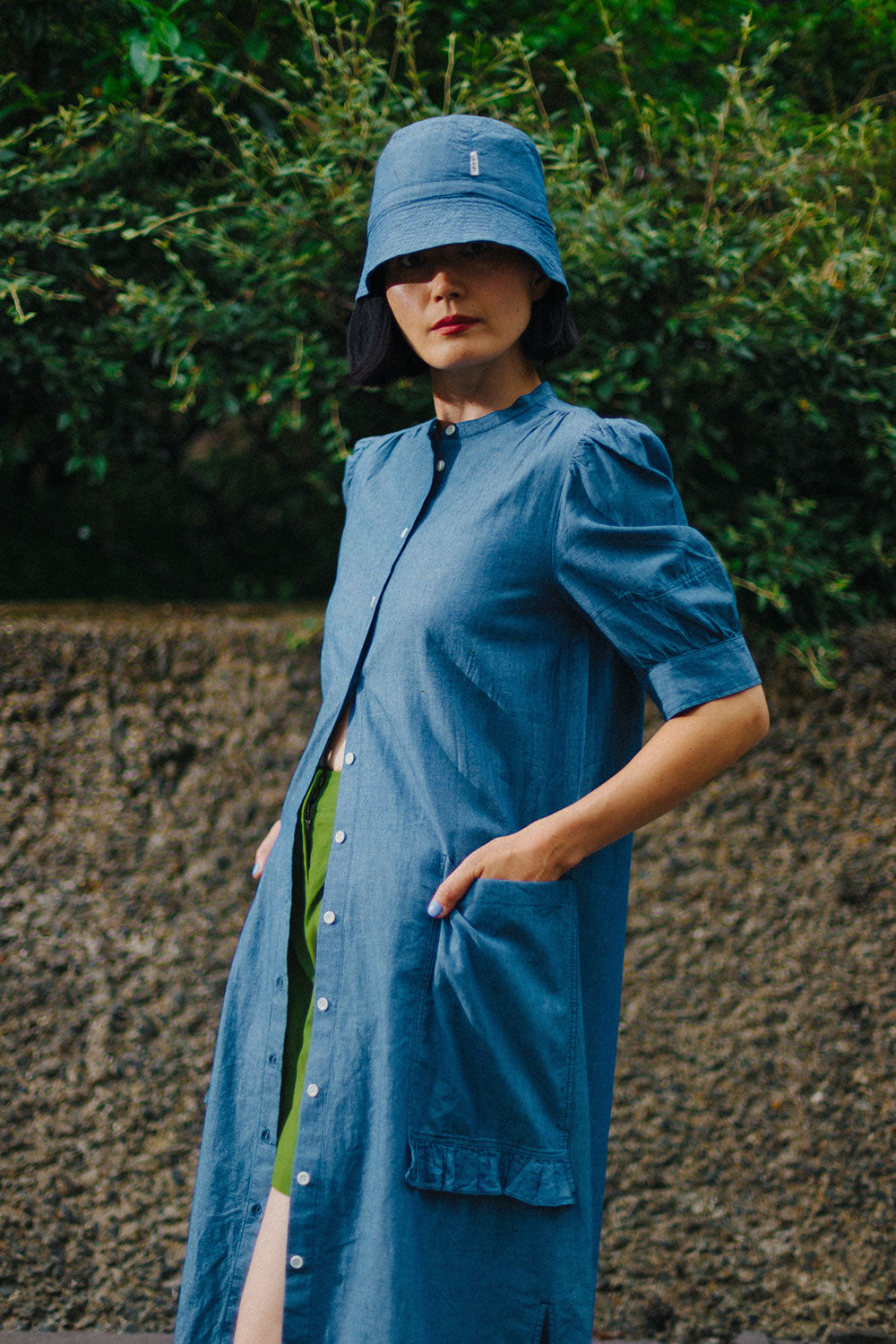 A woman stands in front of green hedges behind a concrete wall, with her hands in her dress pockets. She wears Saywood's lilghtwash Japanese denim Rosa Puff Sleeve Shirtdress, worn open from the waist over Saywood's Bessie A-line Shorts in rich green. Worn with the Reversible Japanese Denim bucket hat on her head, with the lightwash side showing.