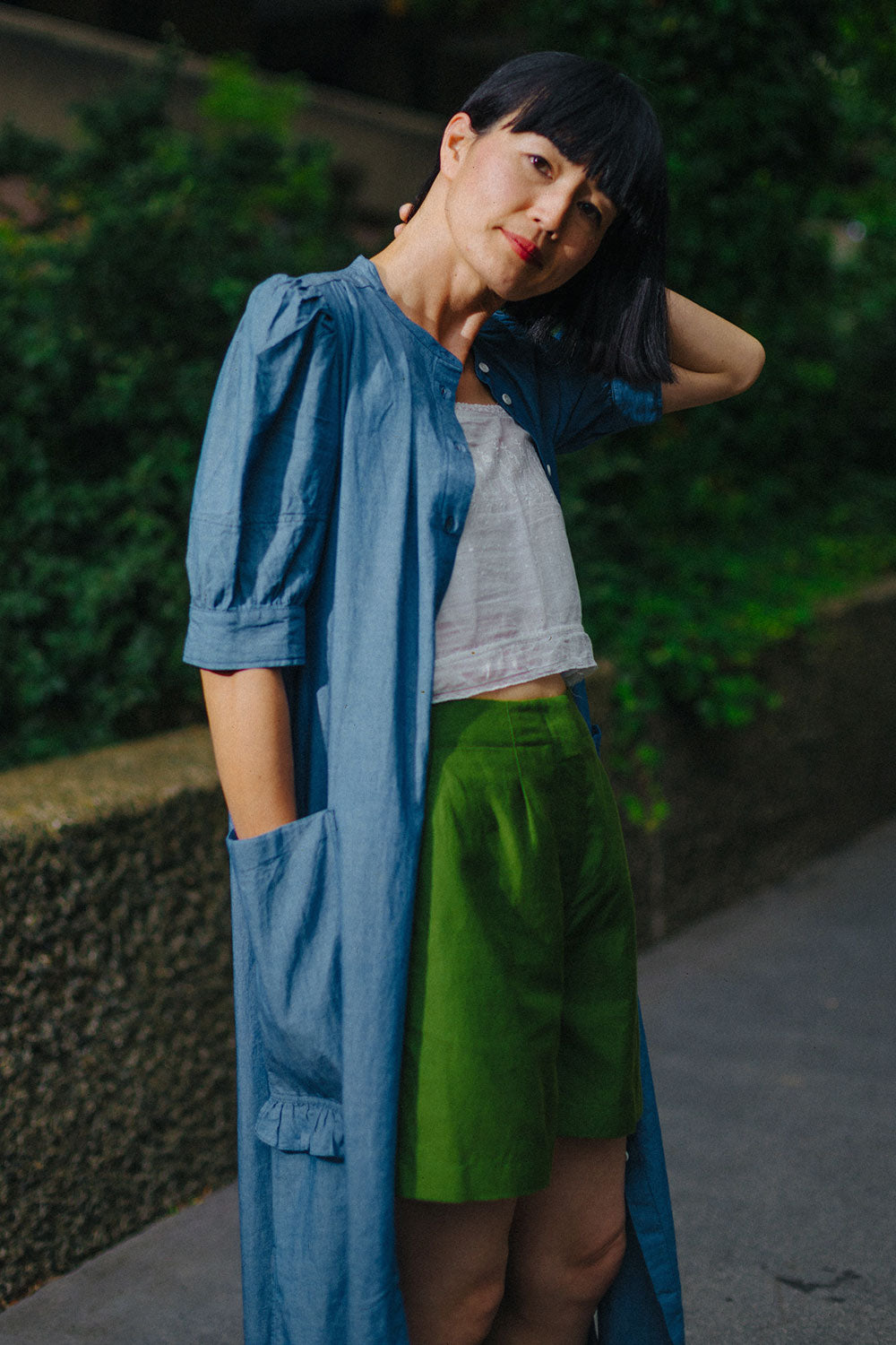 A woman stands in front of green hedges behind a concrete wall, with one hand behind her head moving her hair to one side. She wears Saywood's lilghtwash Japanese denim Rosa Puff Sleeve Shirtdress, worn open over a white vintage cropped vest and Saywood's Bessie A-line Shorts in rich green.
