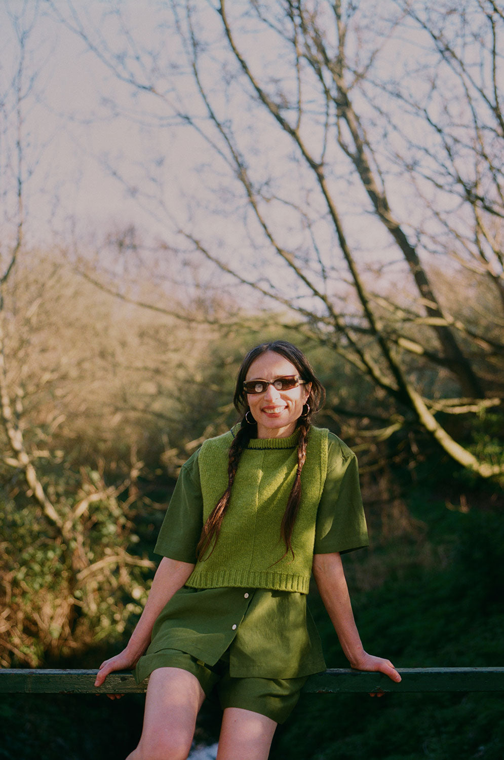 A woman with plaited pigtail hair is in top of green railings on a bridge overlooking trees and a small river. She is wearing Saywood's Frances Unisex Cuban Short Sleeve Shirt in olive green with matching Bessie A-line Shorts. Worn with the new knitted wool sweater vest from Saywood, and sunglasses.