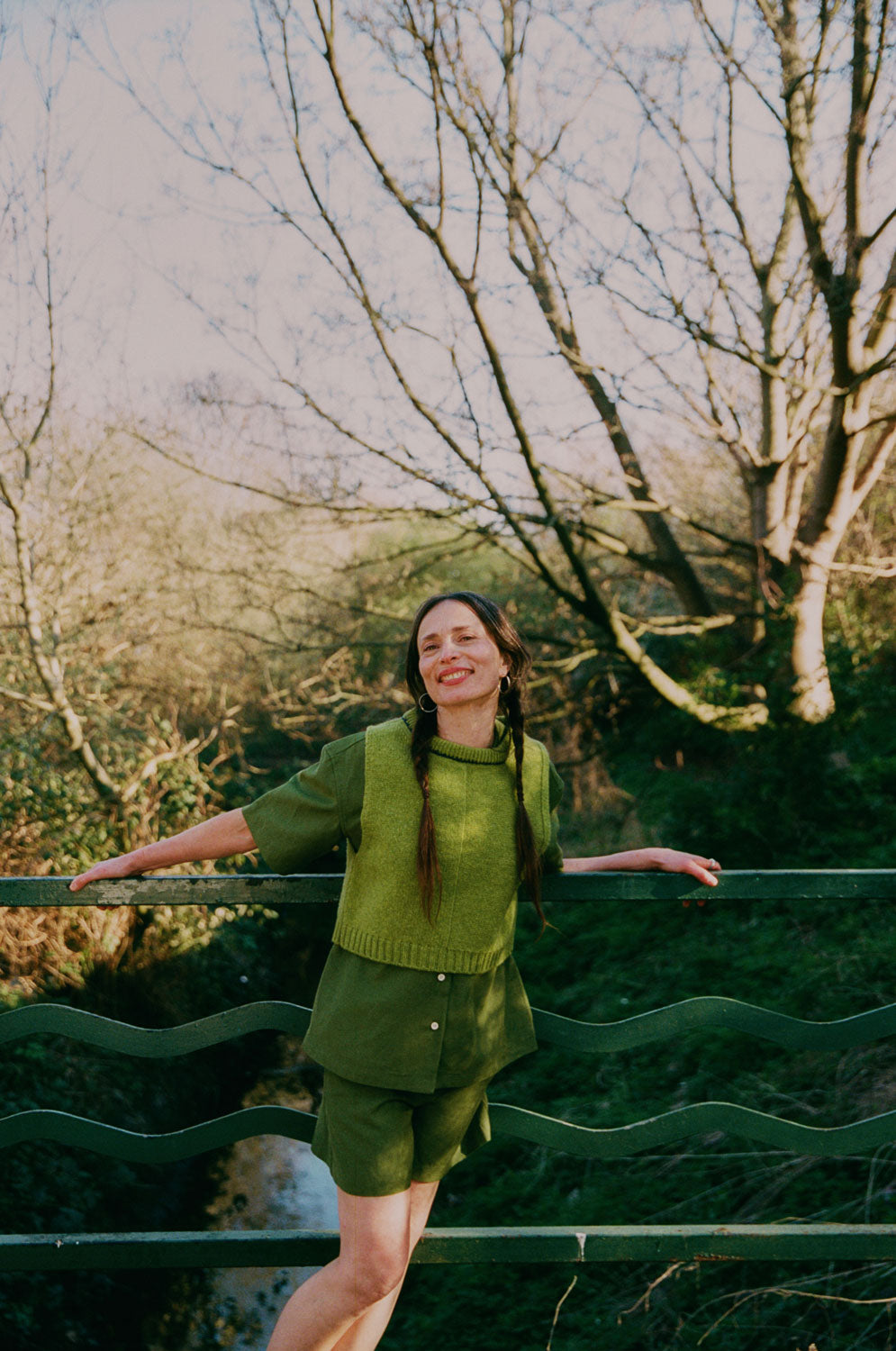 A woman with plaited pigtail hair is standing in front of green railings on a bridge overlooking trees and a small river. She is leaning away from the rails and wearing Saywood's Frances Unisex Cuban Short Sleeve Shirt in olive green with matching Bessie A-line Shorts/ Worn with the new knitted wool sweater vest from Saywood.