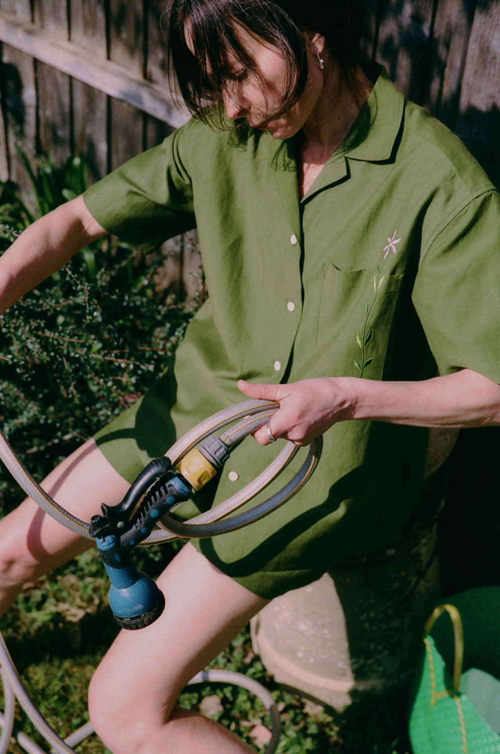A woman is perched on a garden pot, holding a garden hose, with a wooden fence behind her. She is wearing the Frances Unisex Cuban Shirt in olive green, with short sleeves and the signature embroidered flower in lilac on the check pocket. Worn with the Bessie A-line Shorts in olive green as a matching twin set. The image is shot slightly from above, with a green garden basket seen in the corner.
