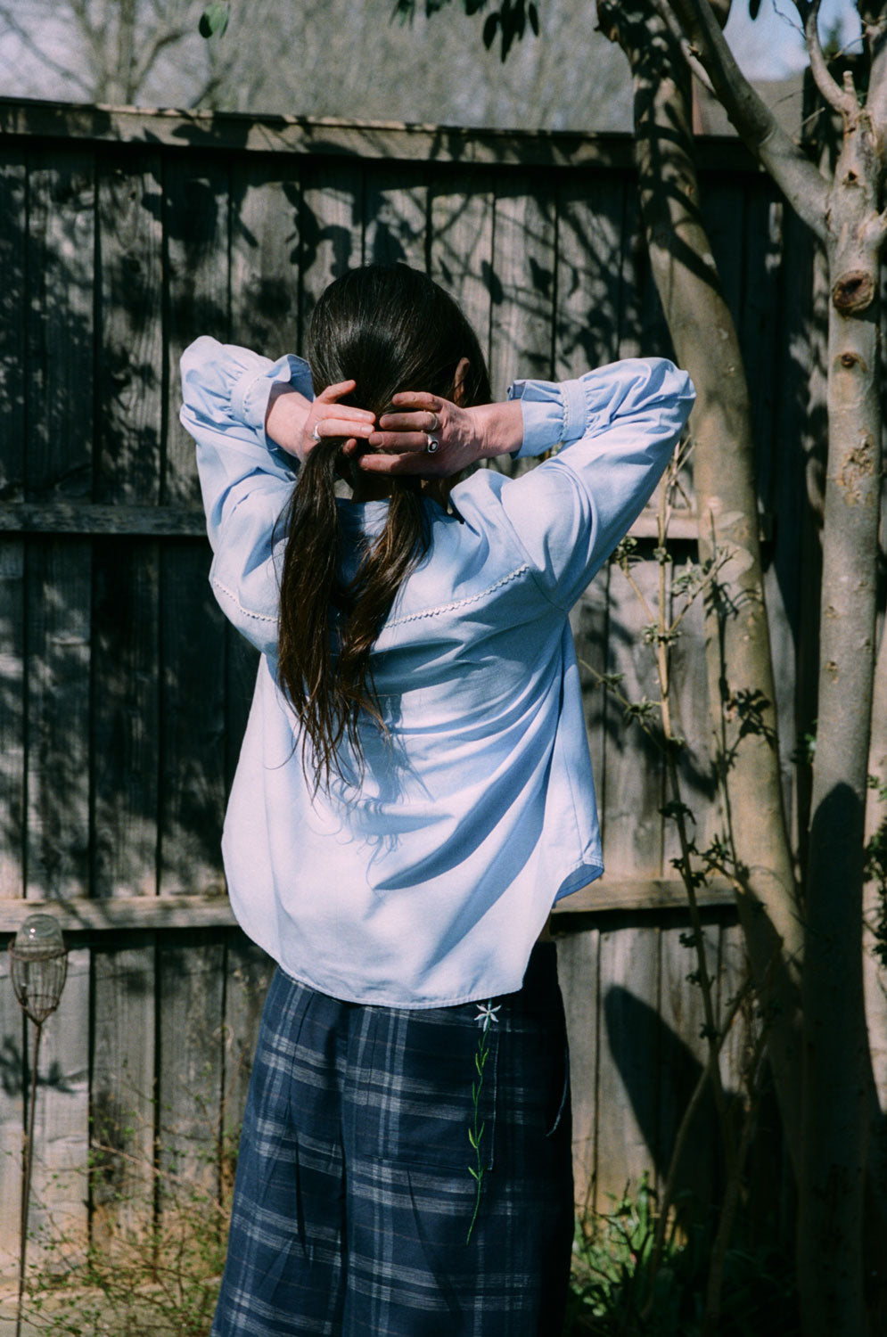 A woman stands with her back to the camera. She is wearing Saywood's Marie A-line Blouse in pale blue recycled cotton, trimmed with a narrow lace trim and gathered frill cuffs. She is plaiting her hair, whilst shadows of the trees can be seen on the fence. On the bottom she wears the navy check Amelia Trousers, with embroidered flower on the back pocket.