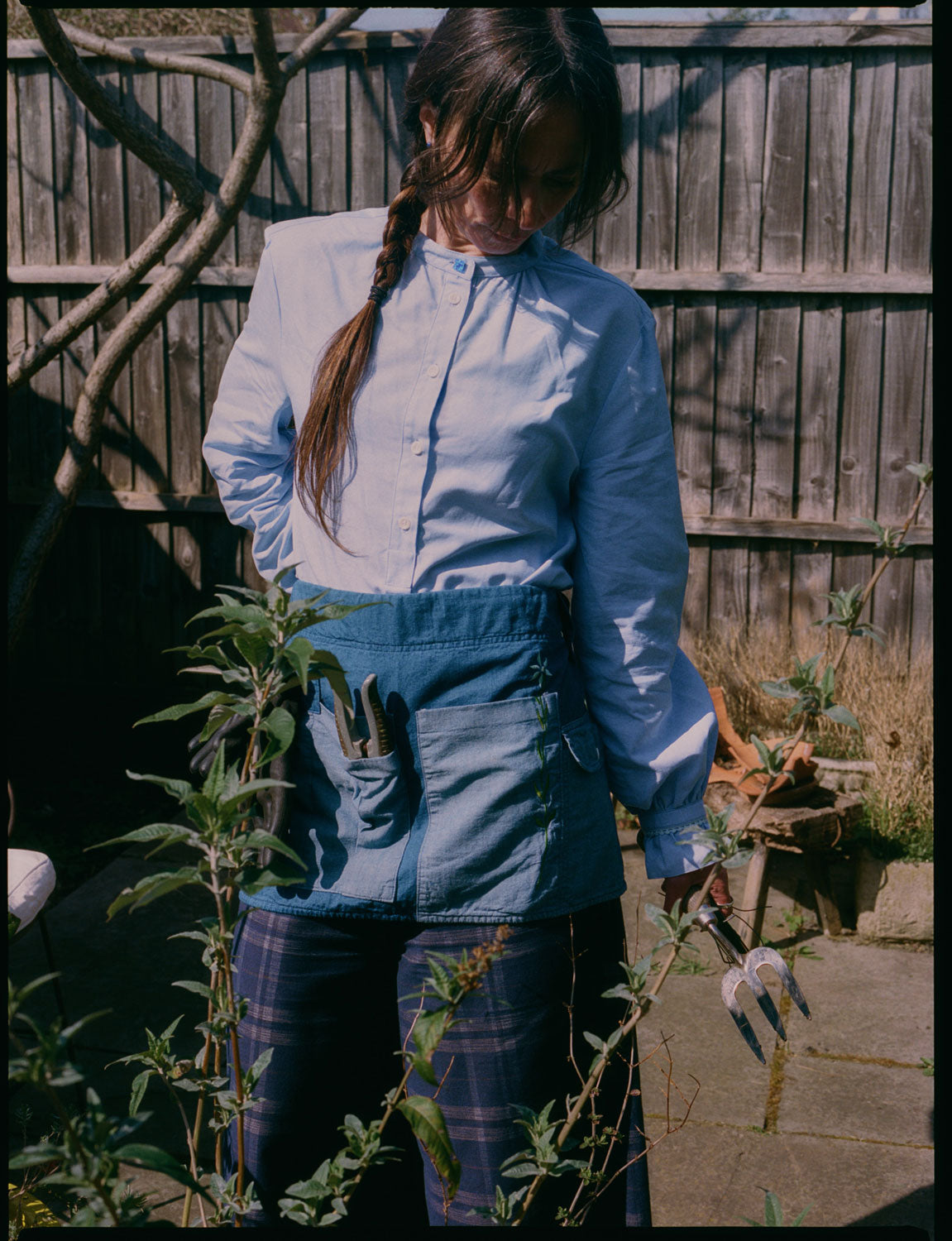 A woman stands in her garden, looking towards the ground, with plants in front of her. She is wearing Saywood's Marie A-line Blouse with gathered neck and cuffs, with a blue vintage glass button at the neckline. Around her waist is the Multifunctional Gardening Tool Belt Apron in Japanese Denim with gardening hand tools in. She wears the navy check Amelia Wide Leg Trousers and is holding a garden fork.