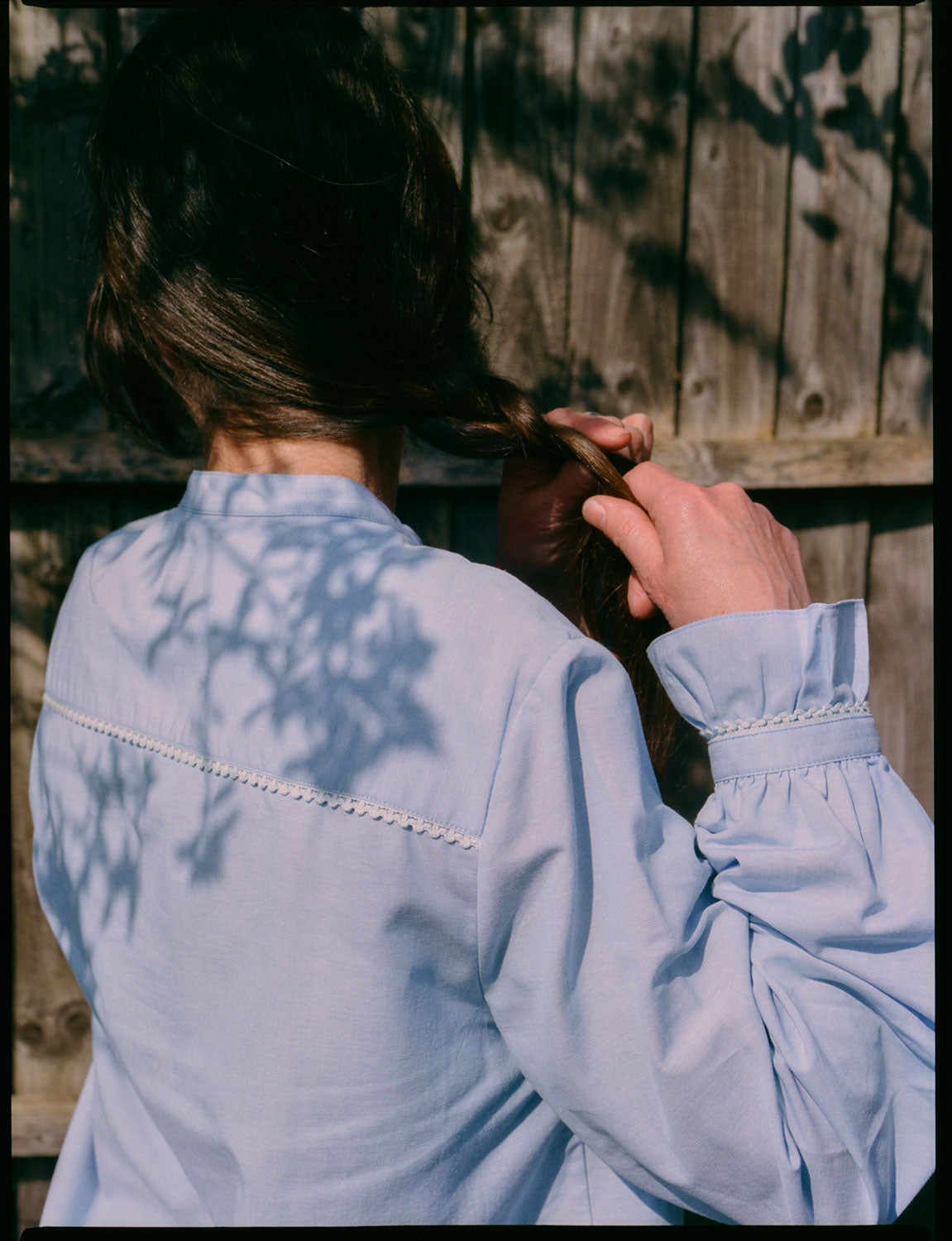 A woman stands with her back to the camera. She is wearing Saywood's Marie A-line Blouse in pale blue recycled cotton, trimmed with a narrow lace trim and gathered frill cuffs. She is plaiting her hair, whilst shadows of the trees are visible on her back.