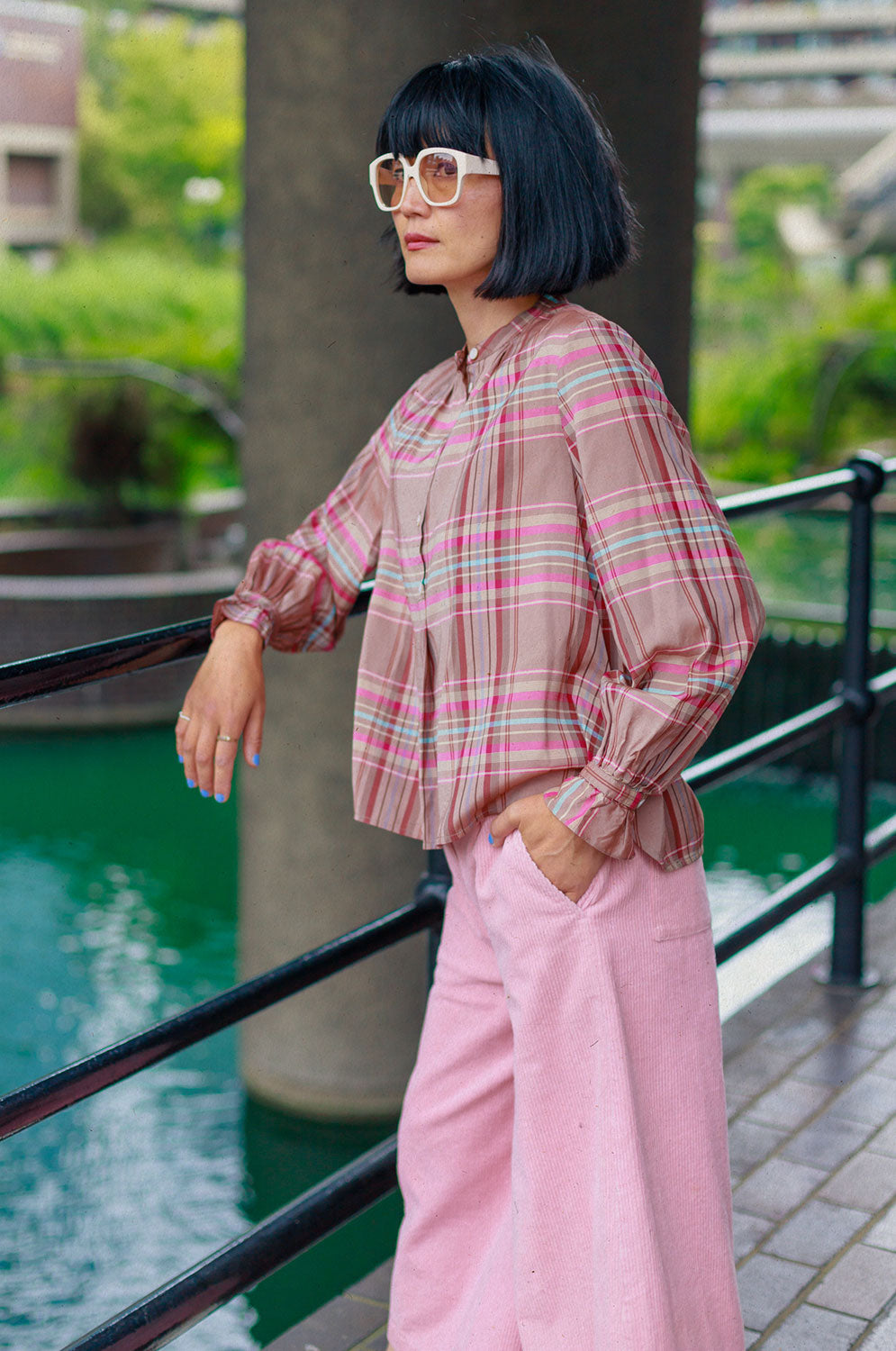 A woman of Japanese heritage is leaning with one arm on an iron railing, overlooking the Barbican pond, with the building just visible in the background. She is wearing Saywood's pink and hazelnut check Marie A-line Blouse with gathered neck details and gathered frill cuffs, with the Amelia Wide Leg Culotte Trousers in pink cotton corduroy. She has one her hand in her pocket, and wears white sunglasses with pink lenses.