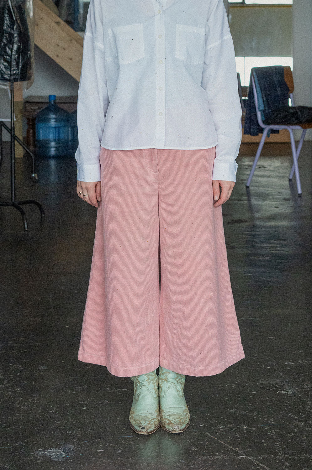 Close up shot of a woman wearing Saywood's Amelia Wide Leg Culotte Trousers in pink cotton corduroy, ankle shy length, with mint green cowboy boots and the Lela Boxy Patch Pocket Shirt in white recycled cotton.