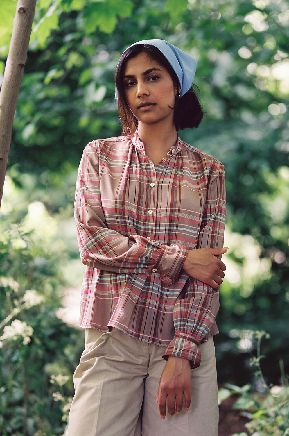 A woman is standing by a tree, with greenery in the background. She is wearing Saywood's Marie A-line Gathered Neck Blouse in pink and hazelnut check, worn with tan chinos and a recycled cotton pale blue bandana in her hair. One arm holds onto her other arms elbow. The gathered frill cuffs can be seen. 