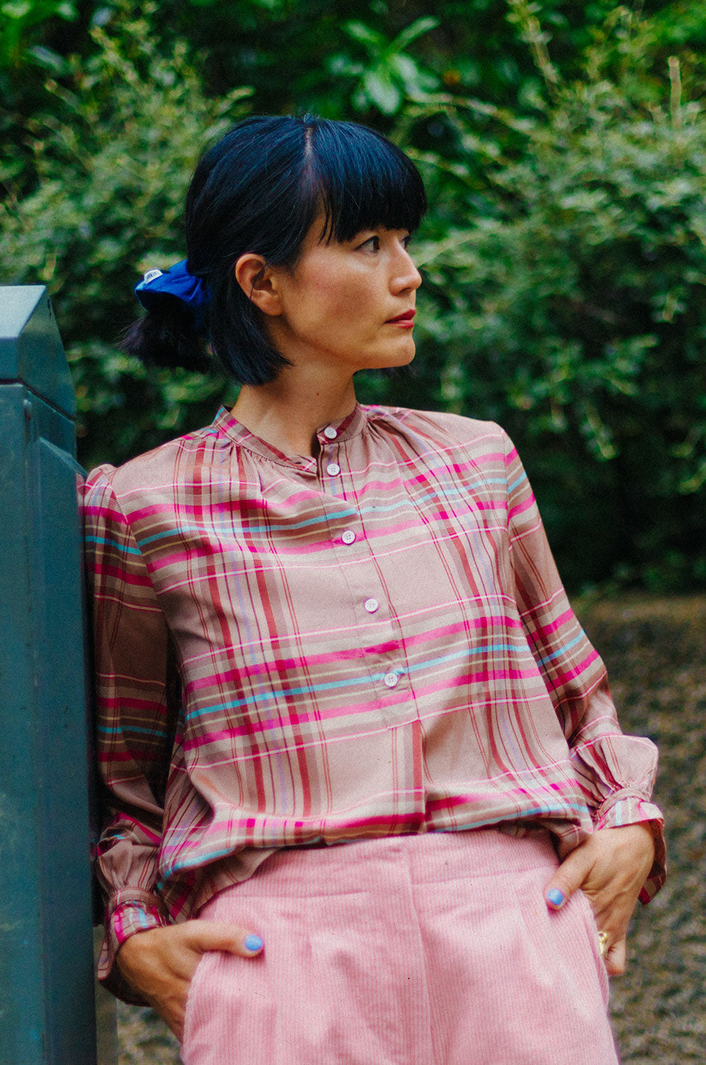 Close up of a woman of Japanese heritage, leaning against a green telephone wire box, with green hedges behind her. She is wearing Saywood's pink and hazelnut check Marie A-line Blouse with gathered neck details and gathered frill cuffs, tucked in at the front to the Amelia Wide Leg Culotte Trousers in pink cotton corduroy. She has her hair half up, tied with the Japanese Denim patchwork scrunchie, with her hands in her pockets.