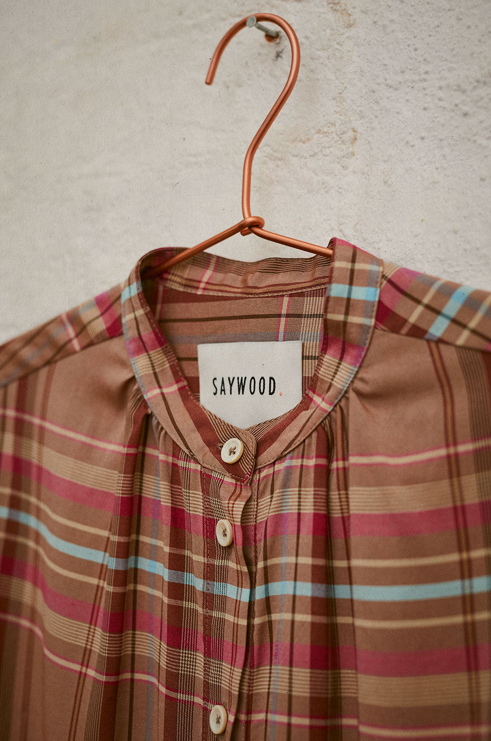 A close up of Saywood's Marie A-line Blouse hanging on a rose gold hanger. the image shoes the gathering at the neckline of the grandad collar, with the ivory 'Saywood' label on the inside back neck.
