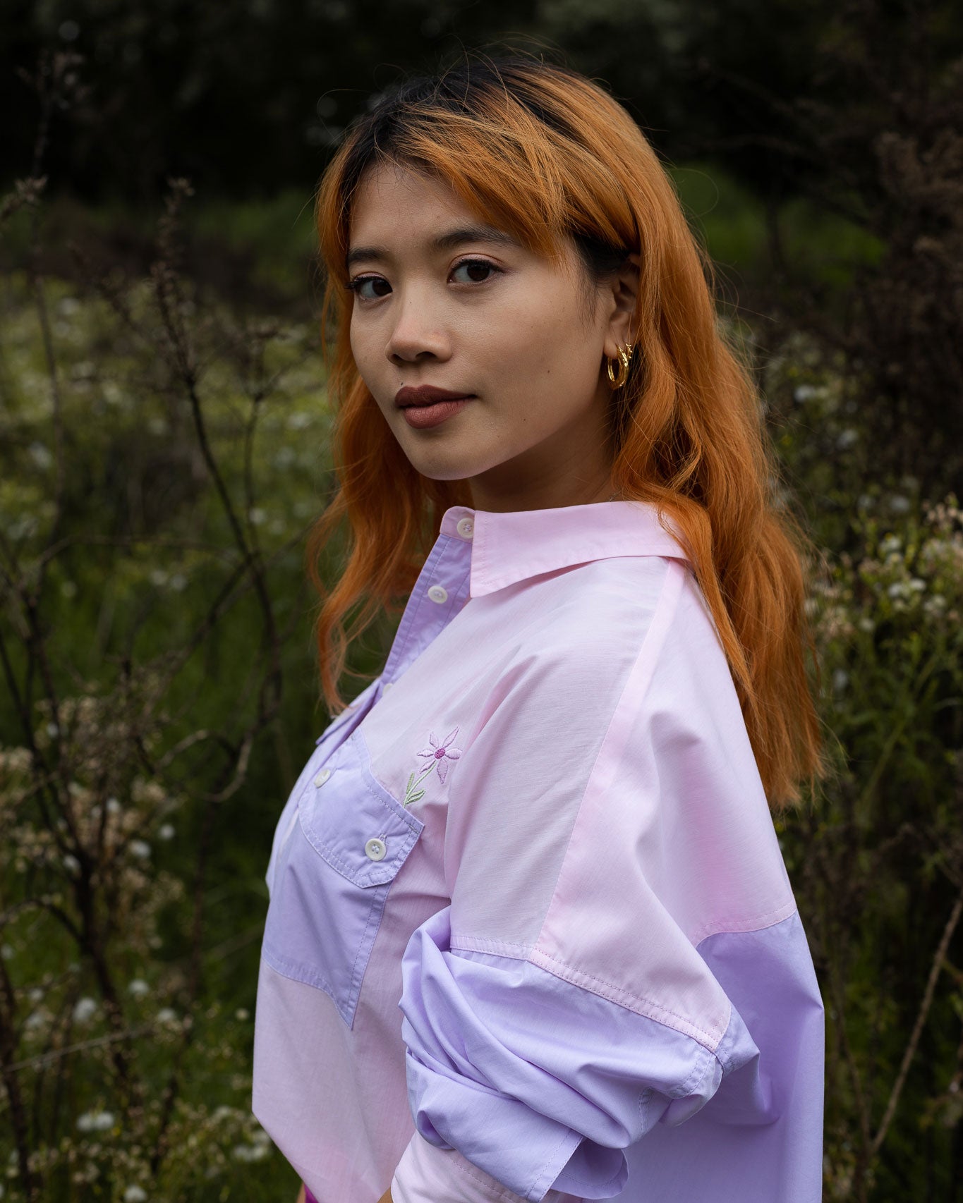 Model stands side on amongst nature, looking at the camera. She wears Saywood's Jules Utility Shirt in lilac and pink colourblock cotton, with the Saywood signature embroidered flower visible just above the patch utility pocket.