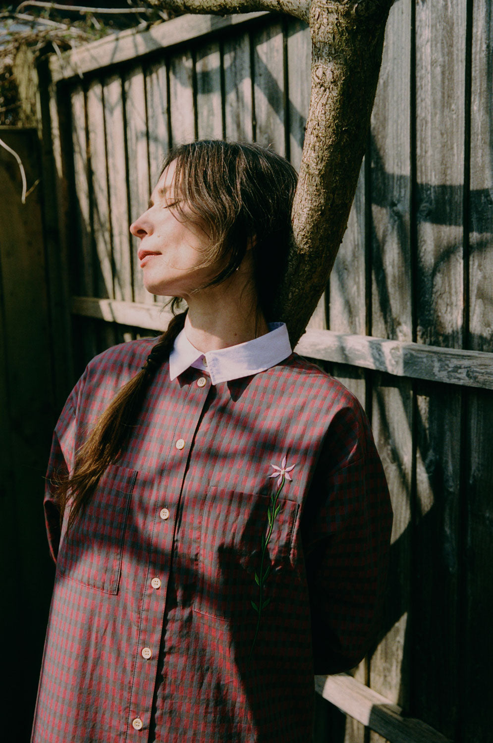 Close up of a woman leaning against a tree in front of a wooden fence. She has her eyes closed and is dappled in sunlight. She wears Saywood's red check Etta Oversized Shirtdress with signature embroidered flower on the pocket, and lilac contrast collar.