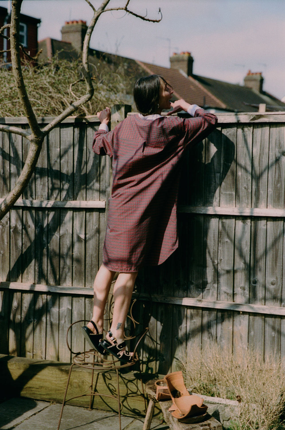 A women is leaning against a fence peeking over the top, with a branchy tree behind her. The image is shot from behind. She wears Saywood's red check Etta Oversized Shirtdress with signature embroidered flower on the pocket, and lilac contrast collar. The box pleat in the back of the dress below the yoke can be seen. She stands on top of a rusty iron decorative chair.