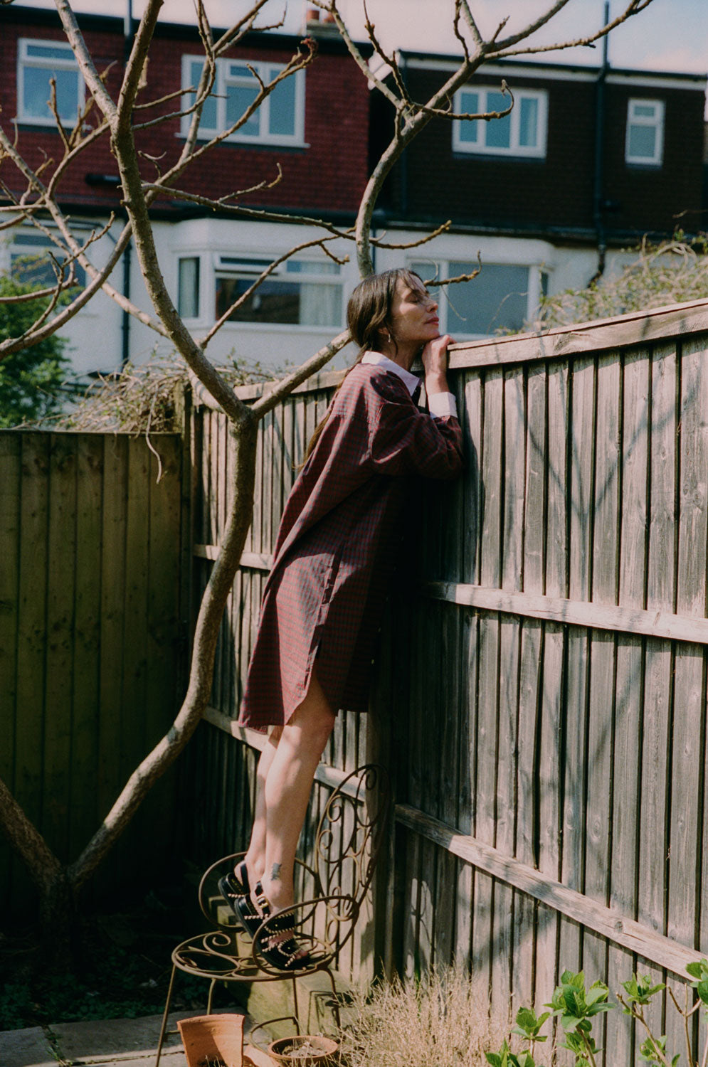 A women is leaning against a fence peeking over the top, with a branchy tree behind her. She wears Saywood's red check Etta Oversized Shirtdress with signature embroidered flower on the pocket, and lilac contrast collar. She stands on top of a rusty iron decorative chair.