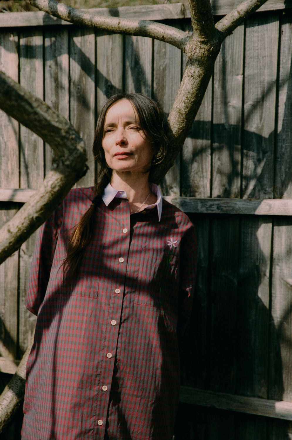 Close up of a woman leaning against a tree in front of a wooden fence. She looks off to the side and is dappled in sunlight. She wears Saywood's red check Etta Oversized Shirtdress with signature embroidered flower on the pocket, and lilac contrast collar.