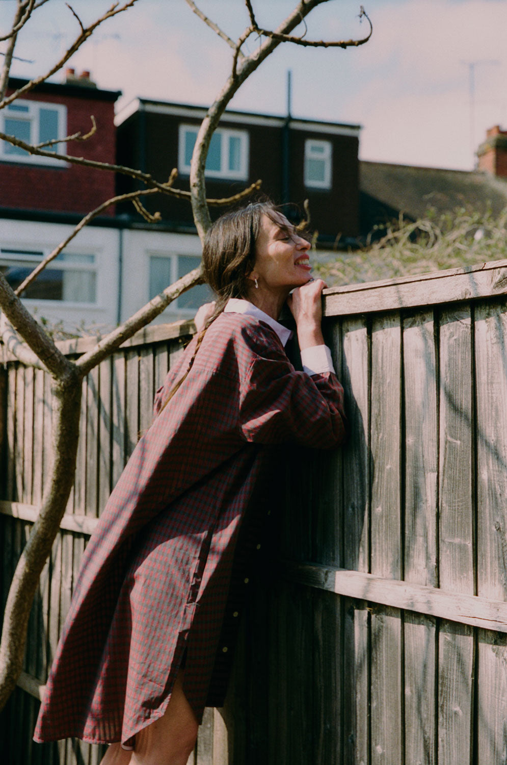 A women is leaning against a fence peeking over the top, with the branches of a tree behind her. She wears Saywood's red check Etta Oversized Shirtdress with signature embroidered flower on the pocket, and lilac contrast collar.