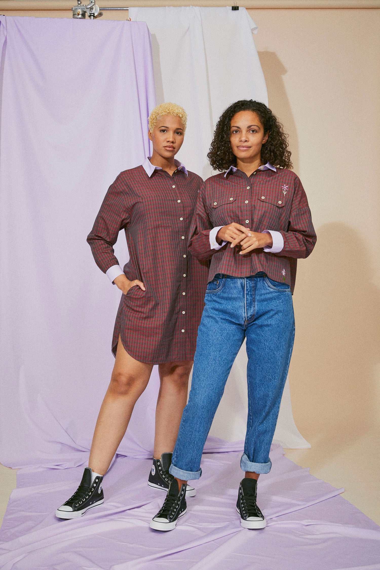Two models stand side by side. The model on the right wears the women's Red Check Shirt, Saywood Studio, Jules Utility Shirt with flower embroidery, Red Check Cotton. The model on the left wears the red check Etta Shirtdress by Saywood