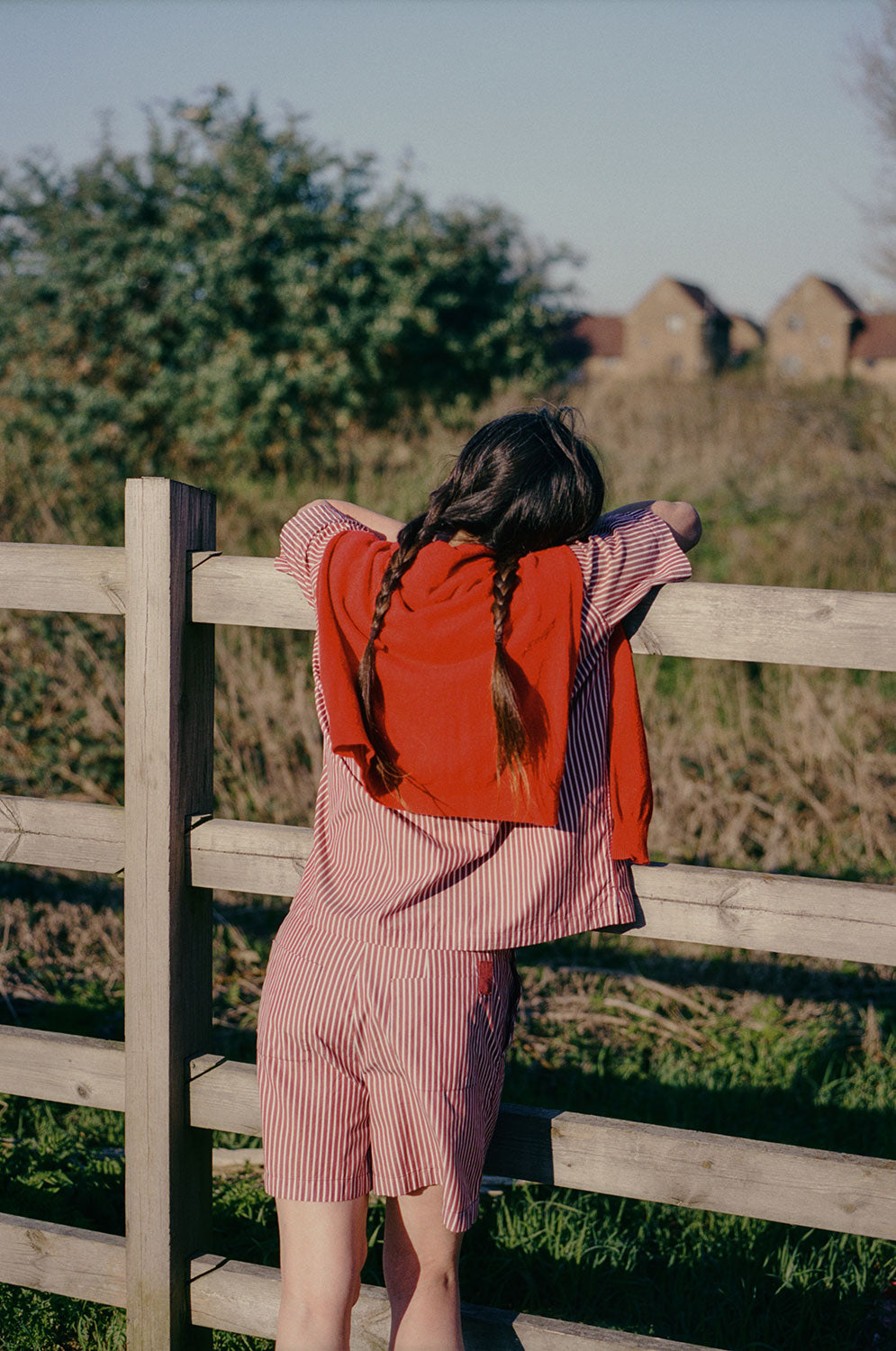 A woman with two plaited pigtails, is leaning on a wooden fence, overlooking grass and houses can be seen far off in the background. She has her back to the camera, and is wearing Saywood's Frances Unisex Cuban Shirt and Bessie A-line Shorts in berry red stripe as a matching set. She has a red cardigan slung round her shoulders.