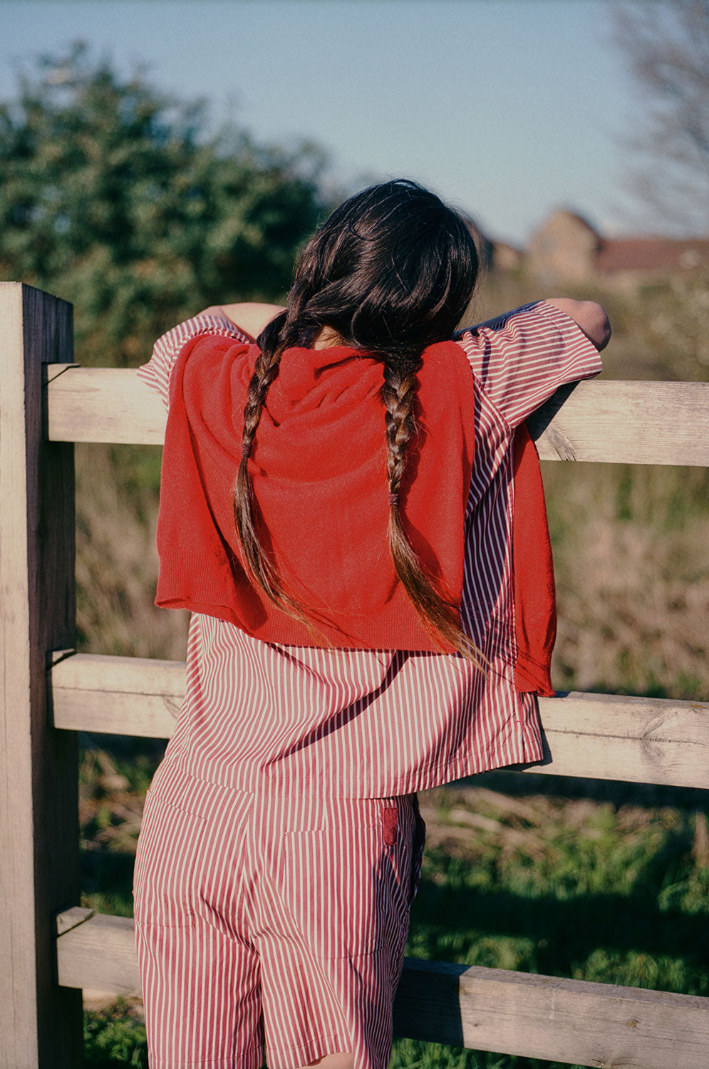 A woman with two plaited pigtails, is leaning on a wooden fence, overlooking grass and houses can be seen far off in the background. She has her back to the camera, and is wearing Saywood's Frances Unisex Cuban Shirt and Bessie A-line Shorts in berry red stripe as a matching set. She has a red cardigan slung round her shoulders. the red herringbone tape detail on the back patch pocket of the Bessie Shorts can be seen.
