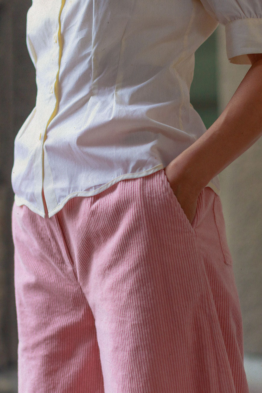 A woman stands sideways to the camera, showing a close up of her mid body. She is wearing Saywood's Joni Puff Sleeve Blouse in lemon. The scallop hem detail and fit and flare darts can be seen clearly. Worn with the Amelia Wide Leg Trousers in pink cotton corduroy. she has her hands in her trouser pockets.