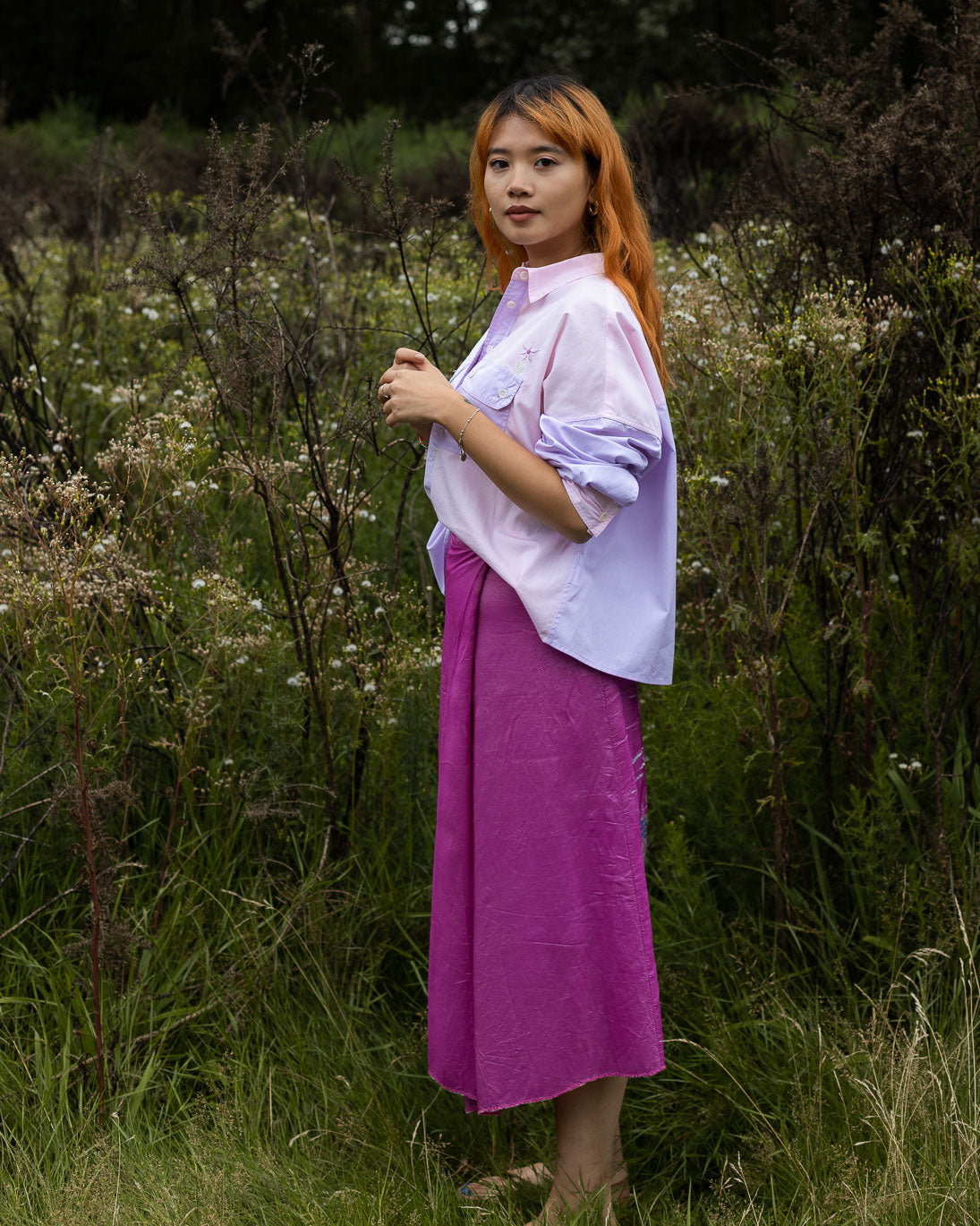 Model stands side on amongst nature, looking at the camera. She wears Saywood's Jules Utility Shirt in lilac and pink colourblock cotton, with the Saywood signature embroidered flower visible just above the patch utility pocket. Worn with sandals and a bright pink wrap skirt.