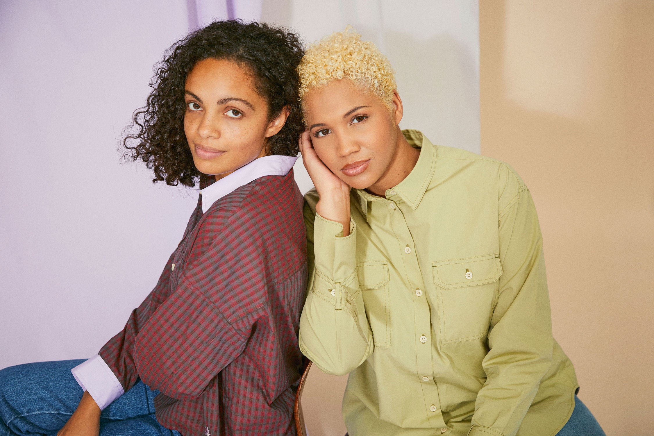 Two women sit, leaning against each other. The woman on the left wears Saywood's Jules Utility Shirt in red check cotton with lilac collar and cuffs. The woman on the right wears the Zadie Boyfriend Shirt in olive.